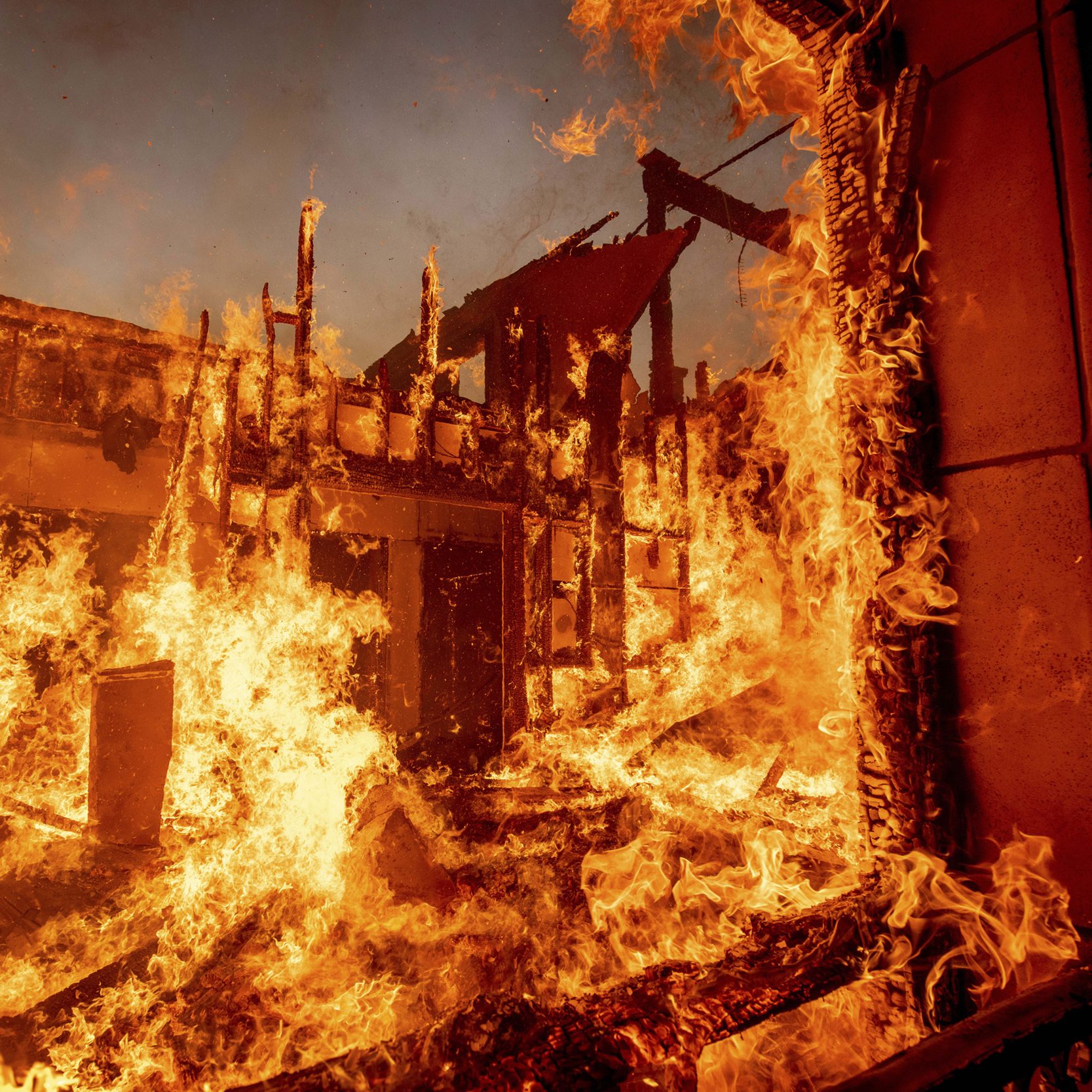 A building engulfed in intense flames, with fire consuming walls and debris, creating a dramatic and chaotic scene against a darker sky background.