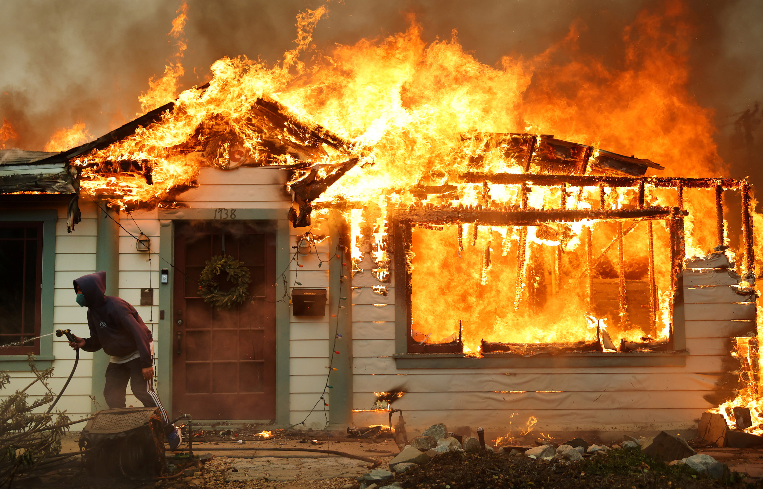 A house is engulfed in flames, with fire blazing through the roof and windows. A person in a hooded sweatshirt moves away, holding a hose.