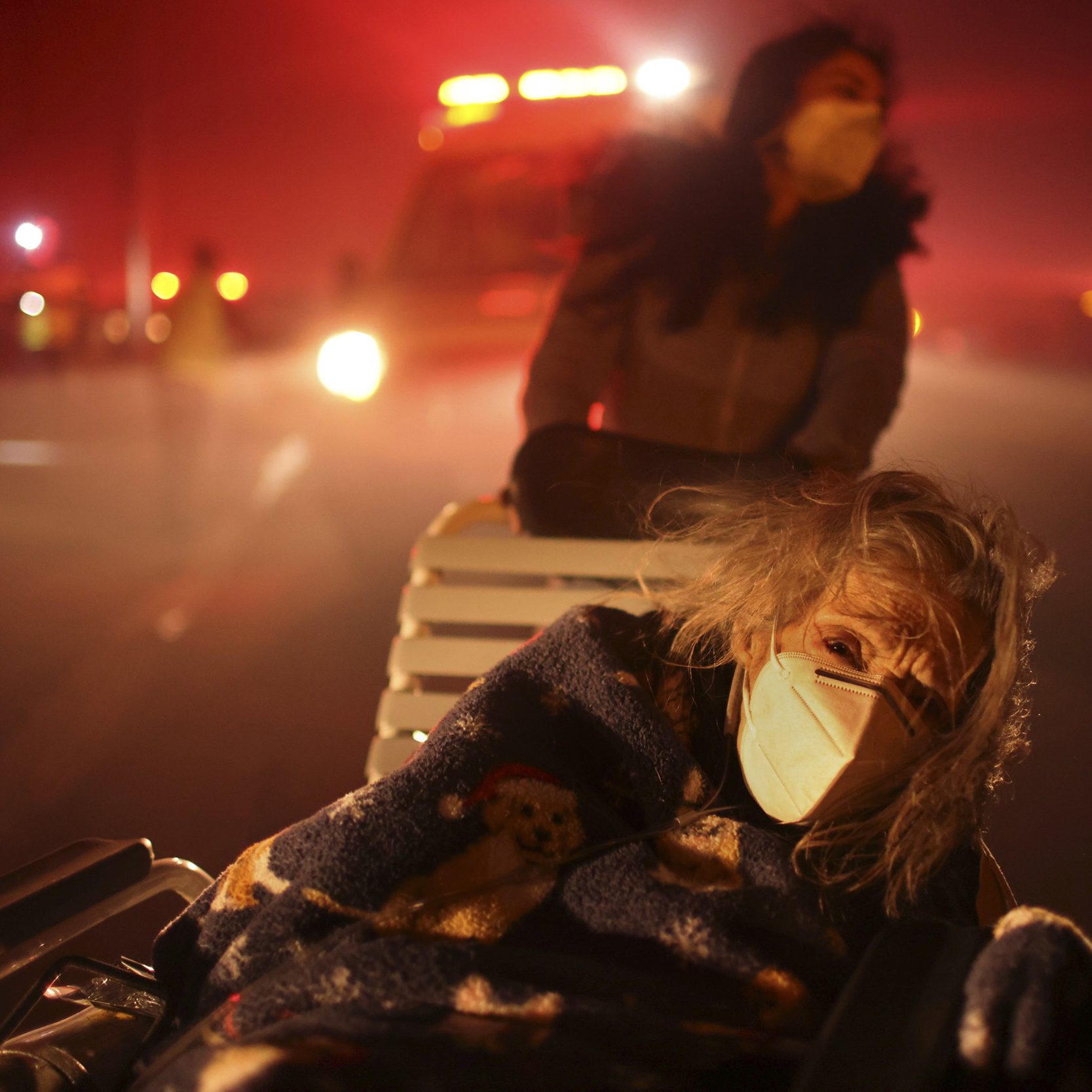 A person in a wheelchair wears a mask and blanket amidst a smoky, reddish scene, with an ambulance and another masked individual in the background.