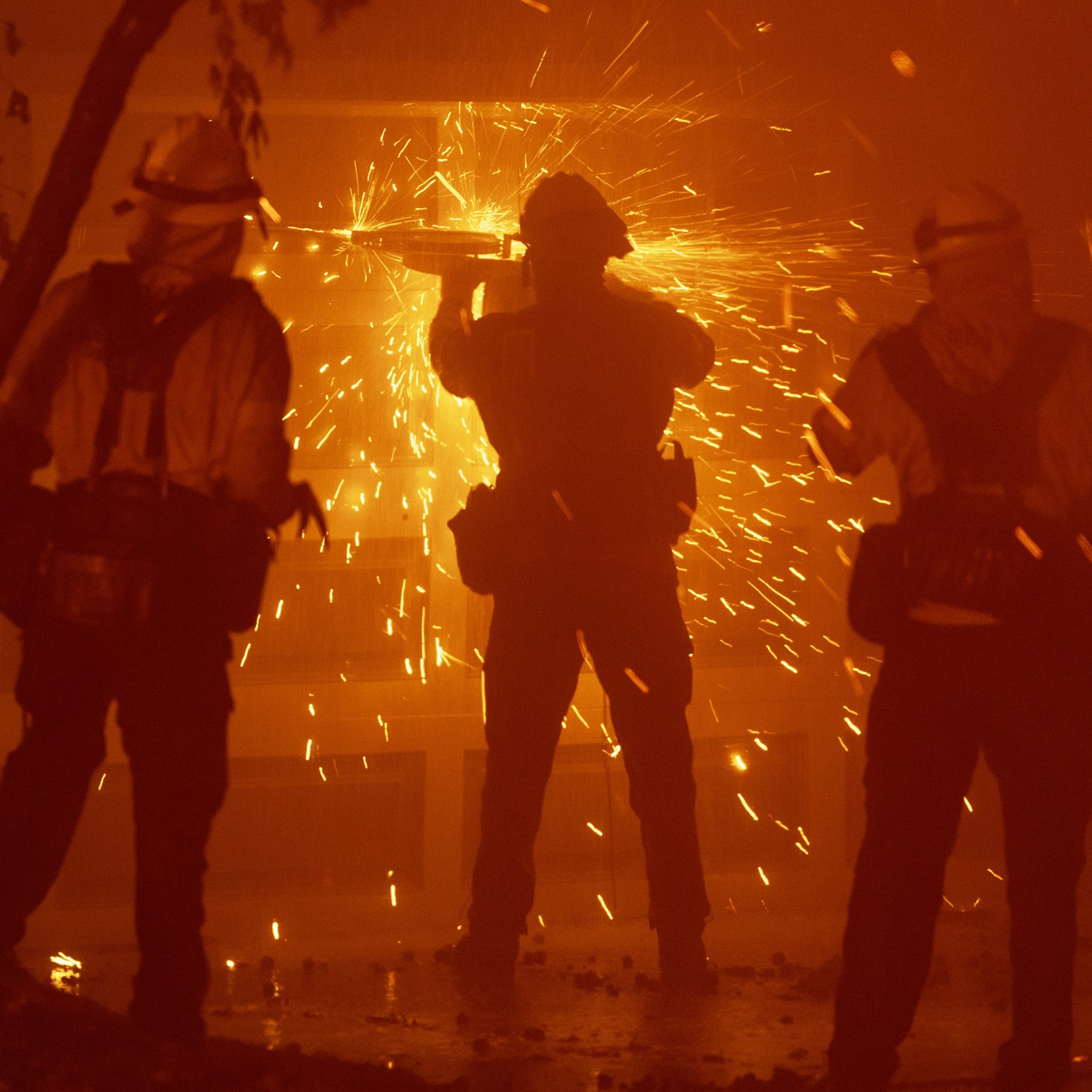 Three firefighters in protective gear stand amid orange flames and smoke, as one uses a tool that creates sparks, illuminating the scene dramatically.