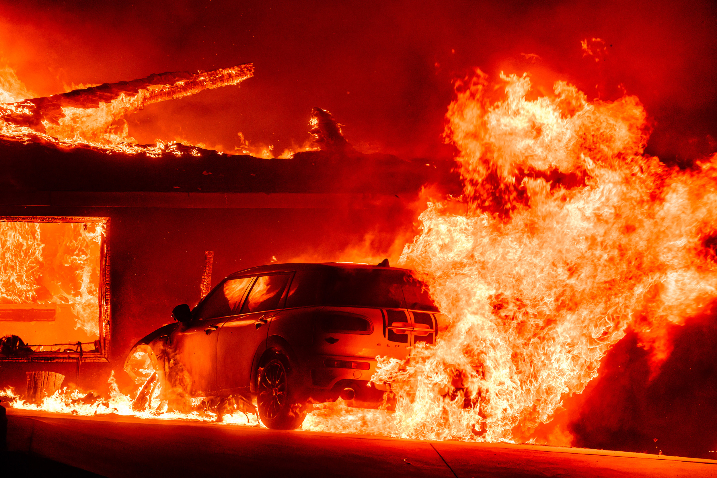 A car is engulfed in bright, intense flames alongside a burning building at night, with firelight reflecting vividly on the vehicle and surrounding area.