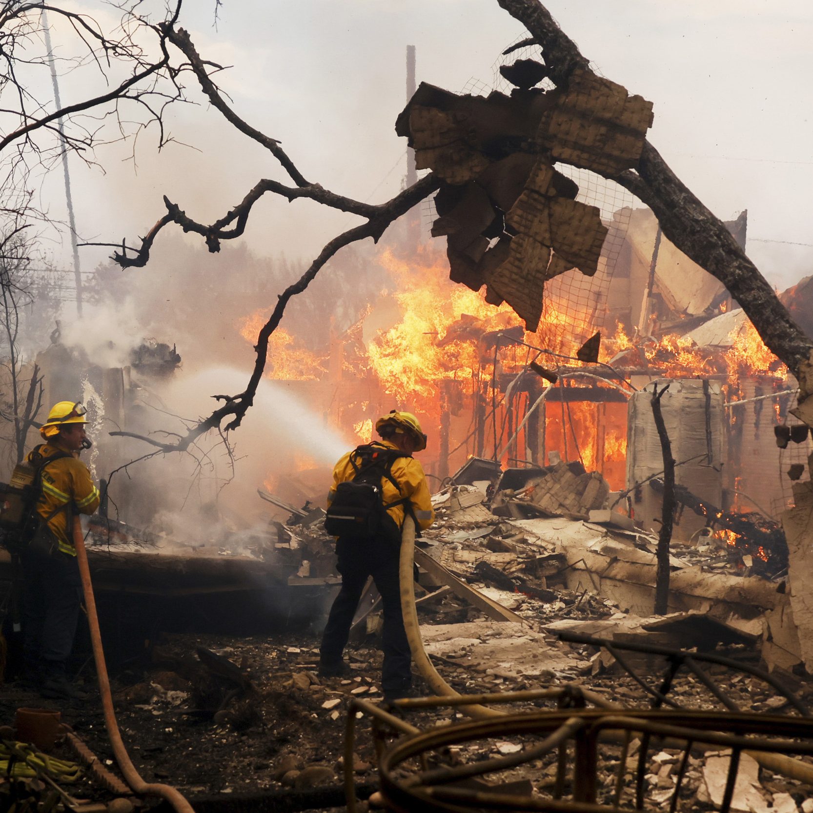 Firefighters in yellow gear are battling a large blaze engulfing a building, surrounded by debris and charred trees, using hoses to spray water.