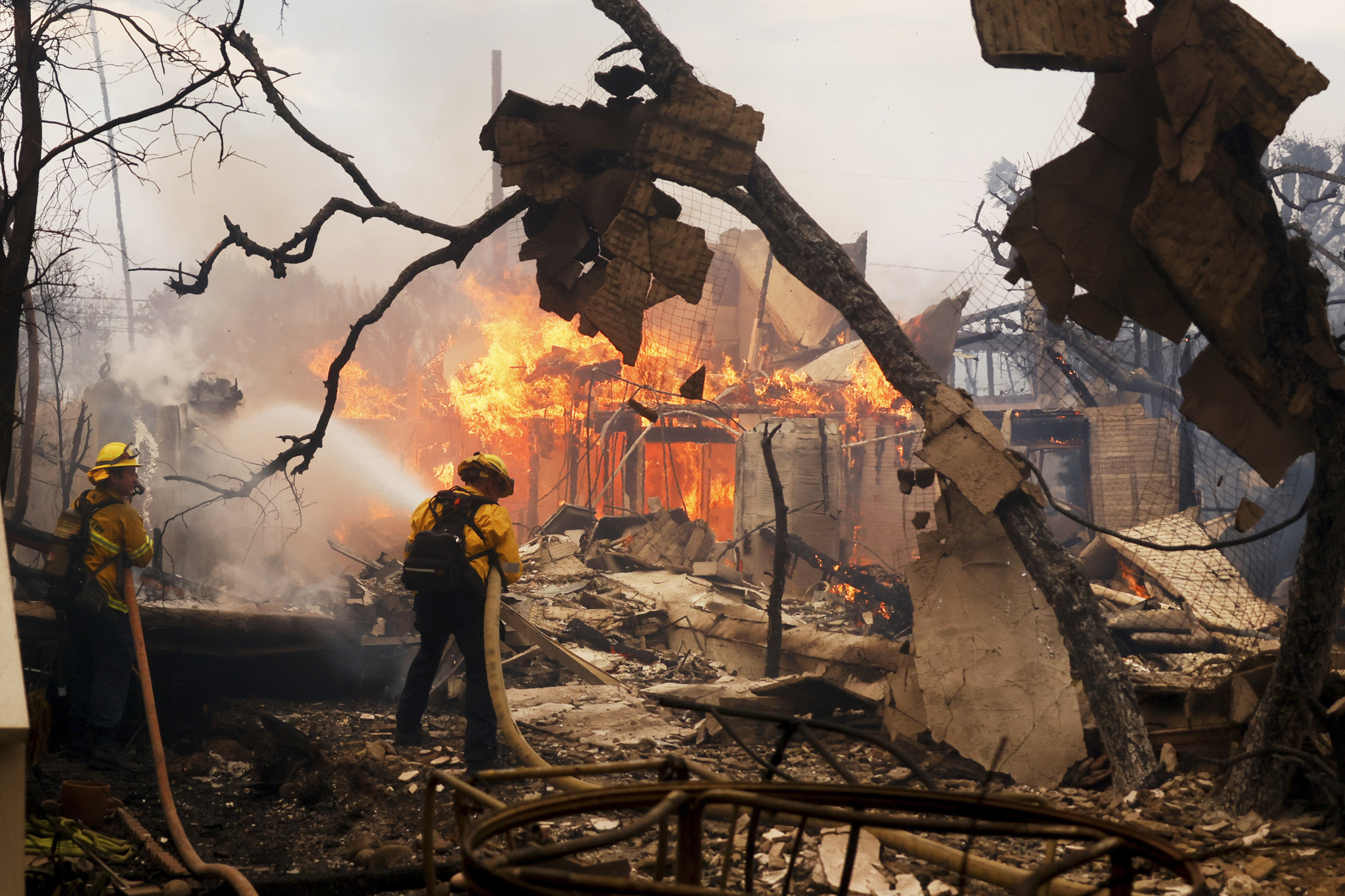 Firefighters in yellow gear are battling a large blaze engulfing a building, surrounded by debris and charred trees, using hoses to spray water.