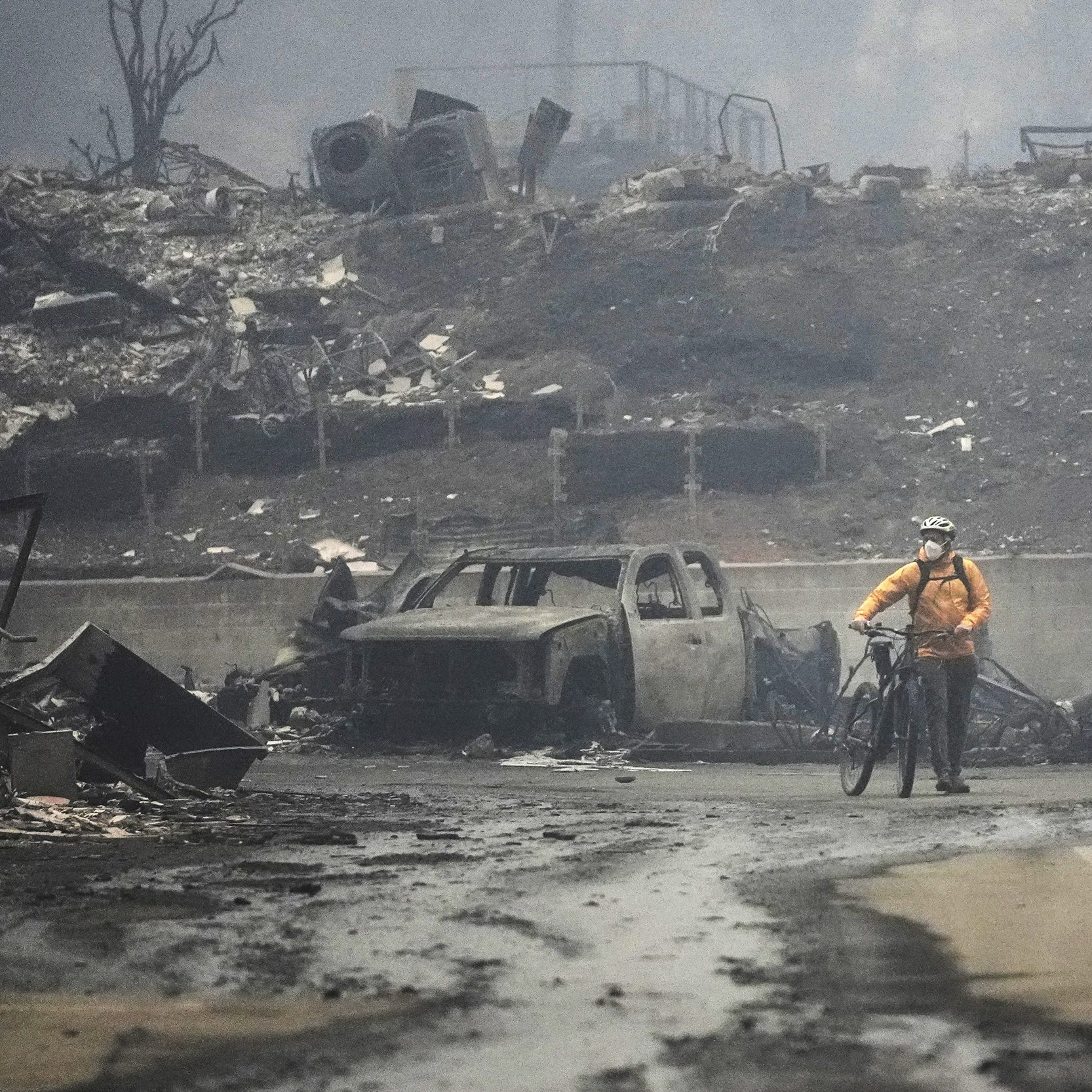 A person in a yellow jacket and helmet walks a bicycle through a bleak, burnt landscape with a charred vehicle and debris scattered around.