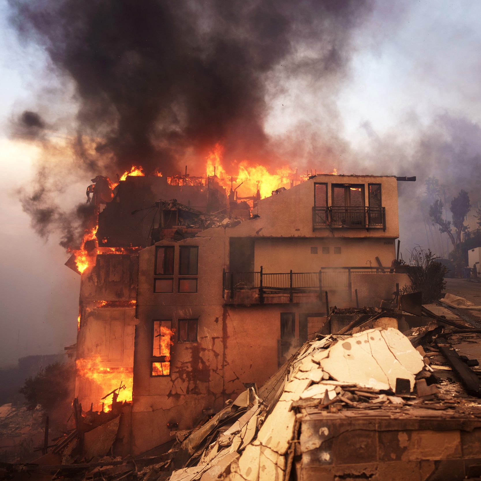 A building is engulfed in flames with thick black smoke rising, while debris is scattered on the ground and the sky is hazy from the smoke.