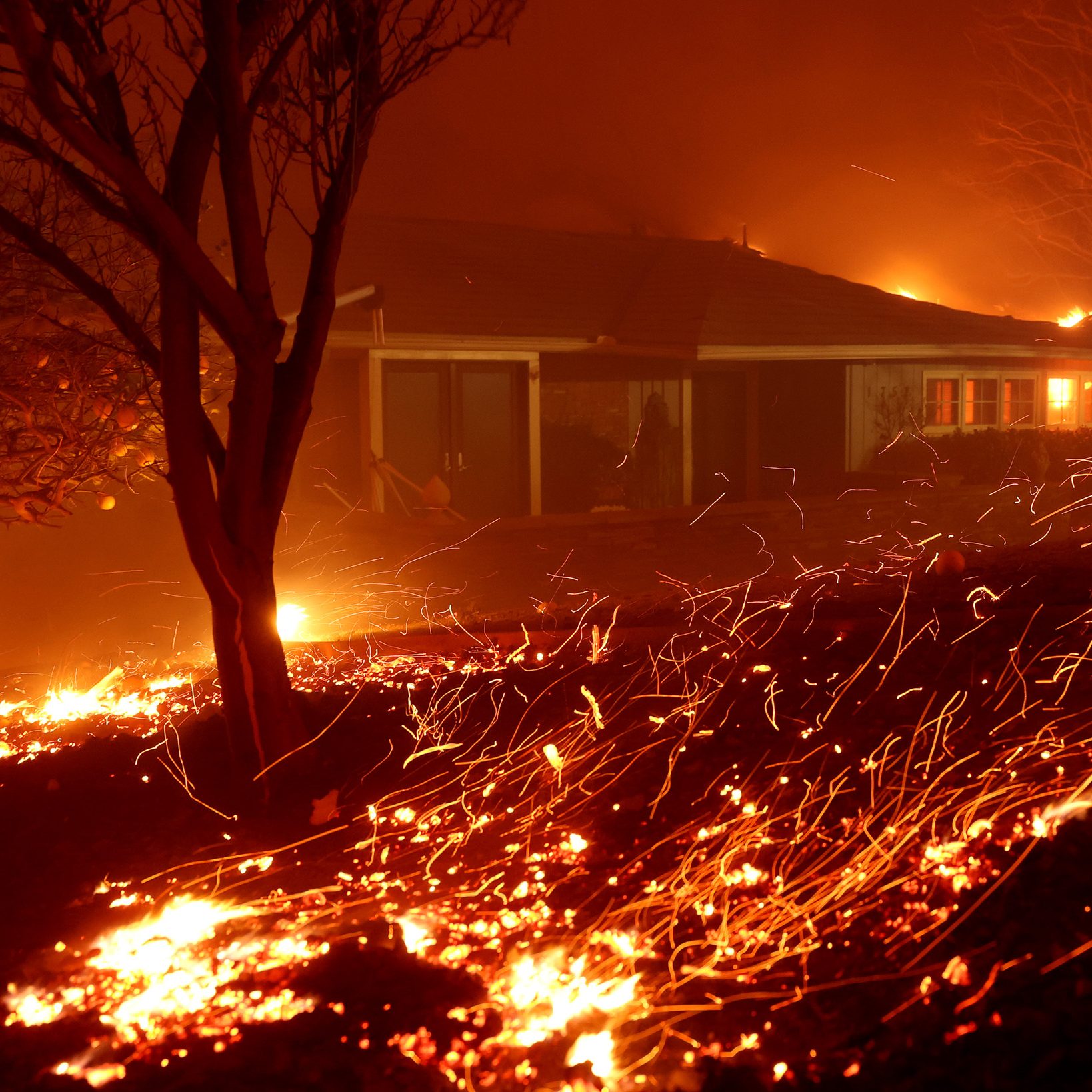 A house is surrounded by flames and smoke in the night. Sparks fly through the dark air, and nearby trees are silhouetted against the fiery backdrop.