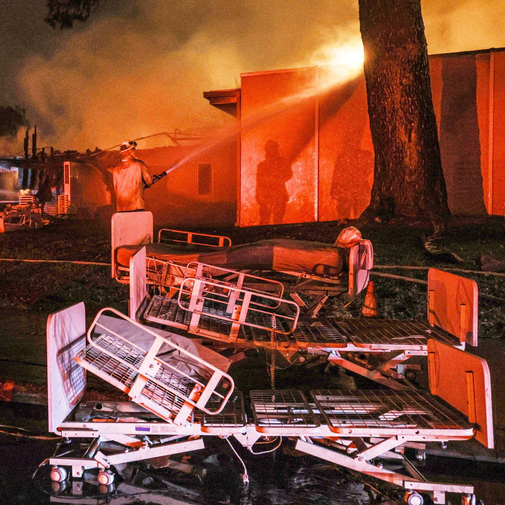 A firefighter is spraying water on a burning building at night. Hospital beds are scattered on the ground, surrounded by orange flames and thick smoke.