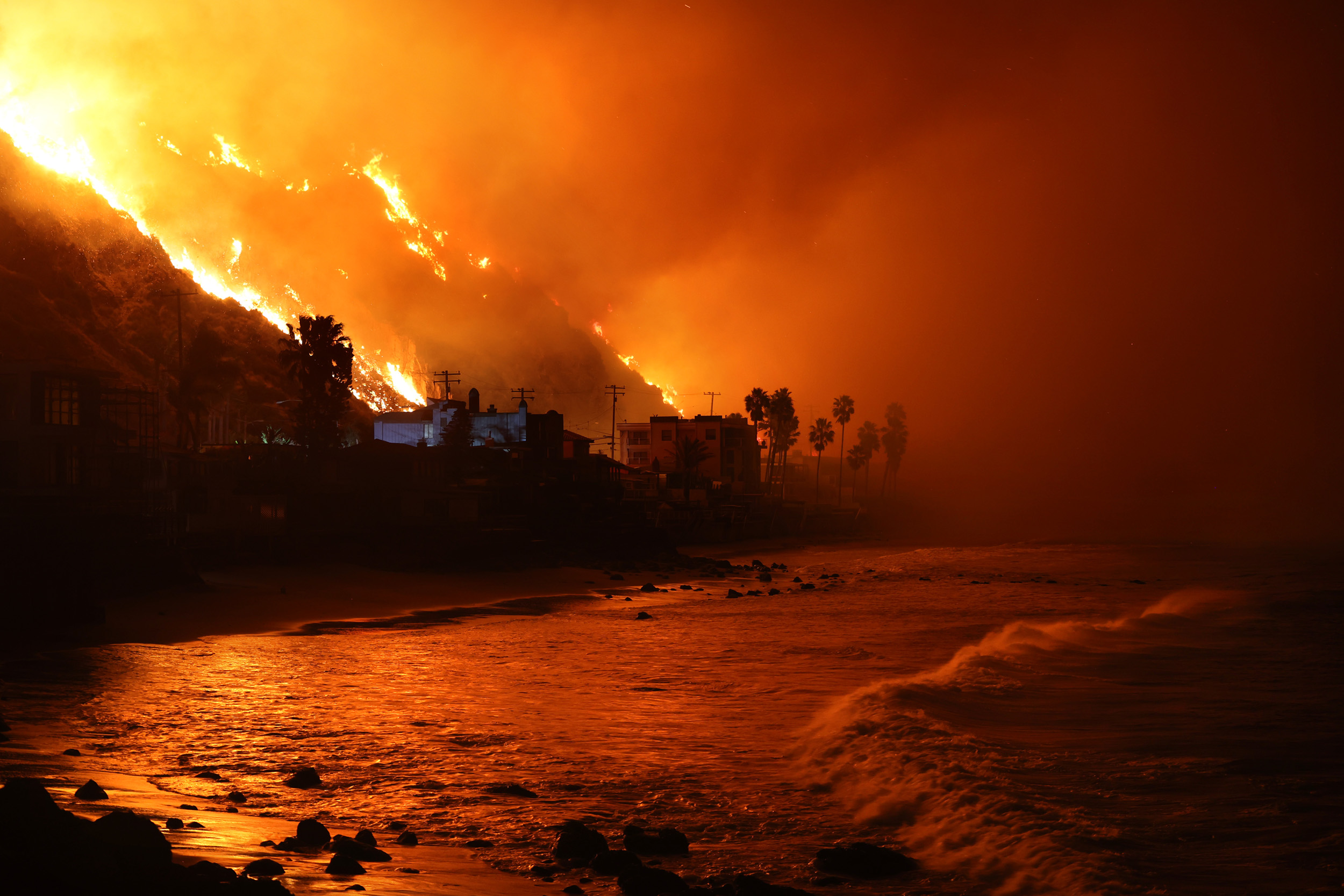 A massive wildfire engulfs a hillside, casting an orange glow over silhouetted buildings and palm trees by the ocean as waves break on the dark shore.