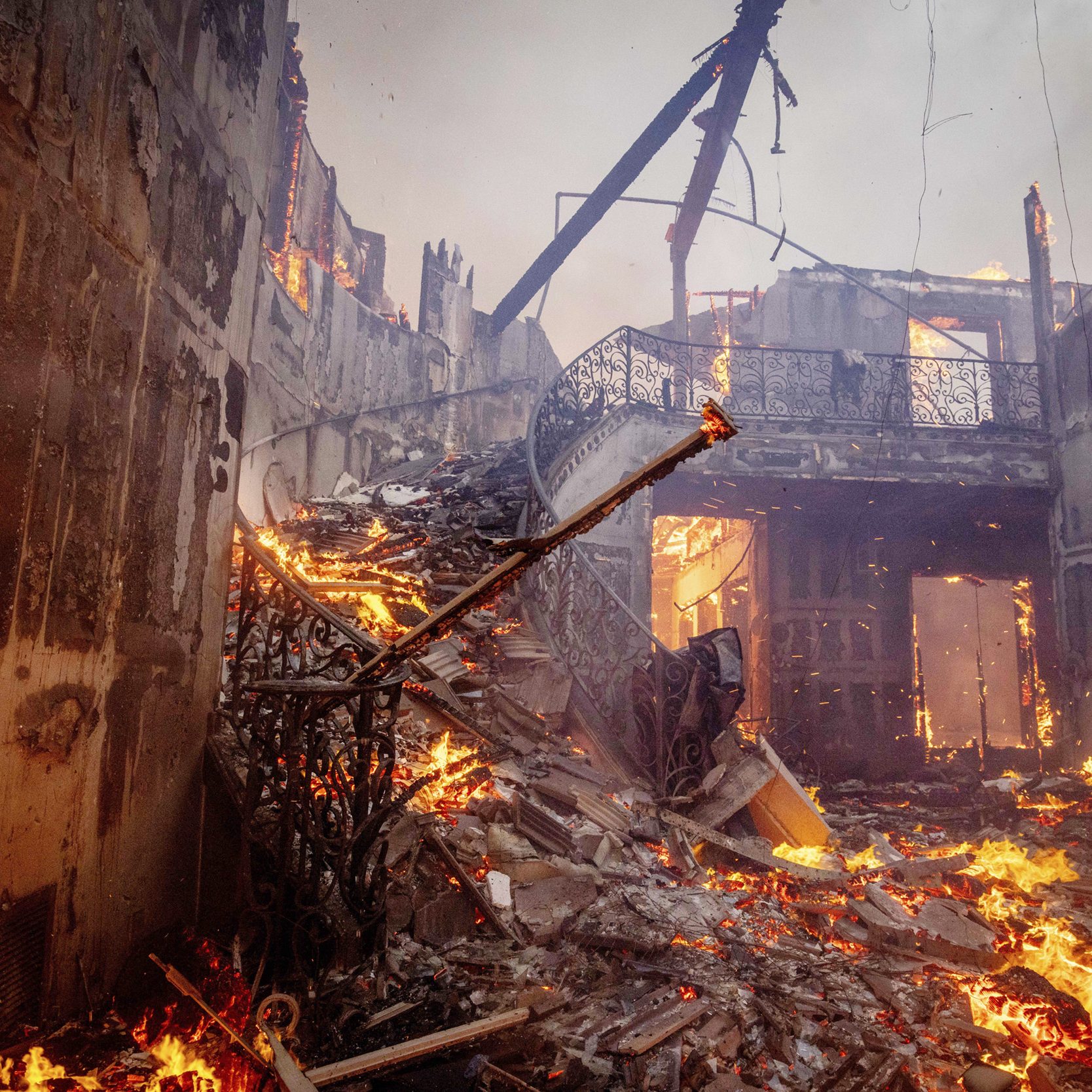 A large, destroyed indoor structure is engulfed in flames, with debris scattered across the floor and charred remains of walls and railings visible.