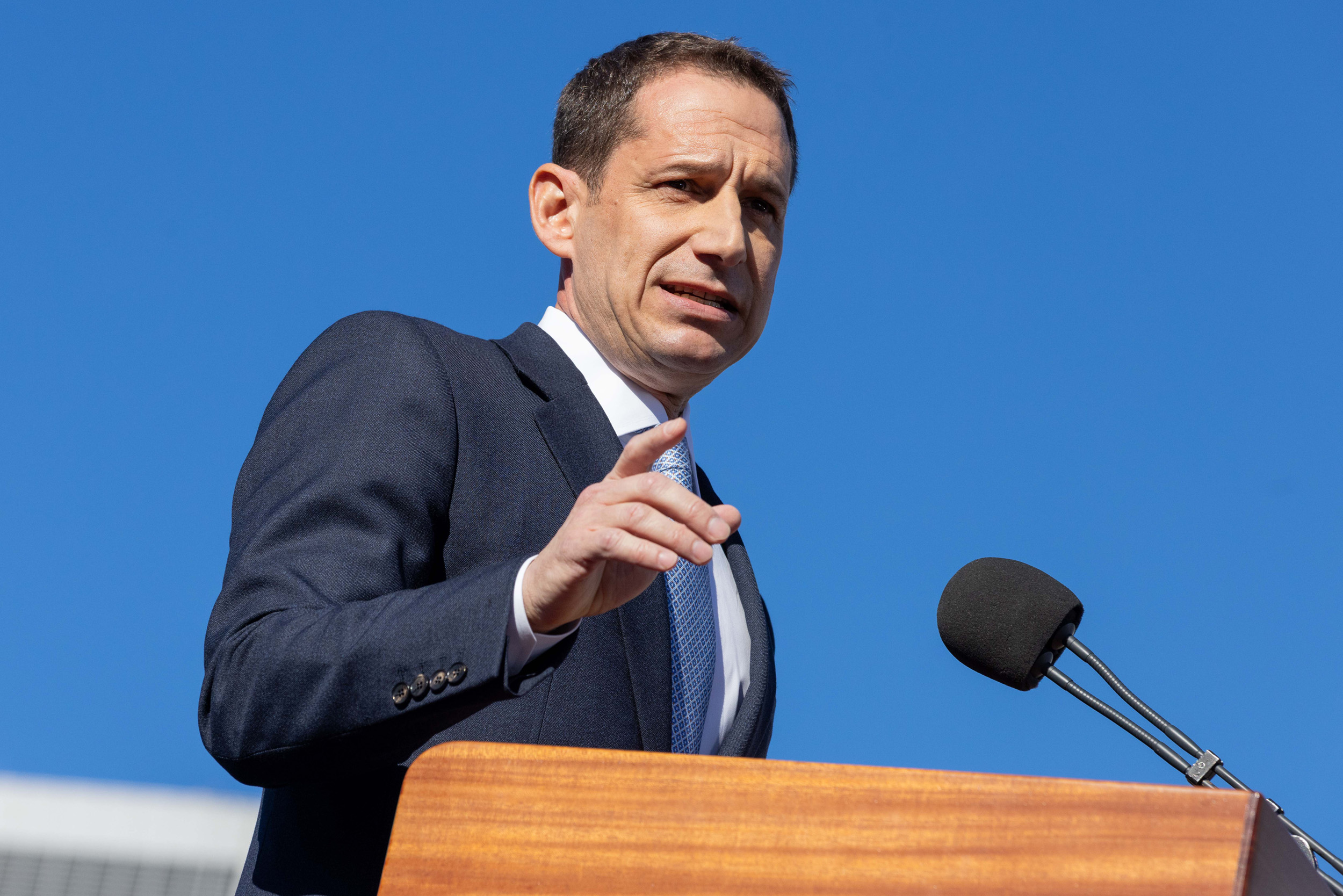 A man in a suit gestures while speaking at a podium with microphones, set against a clear blue sky.