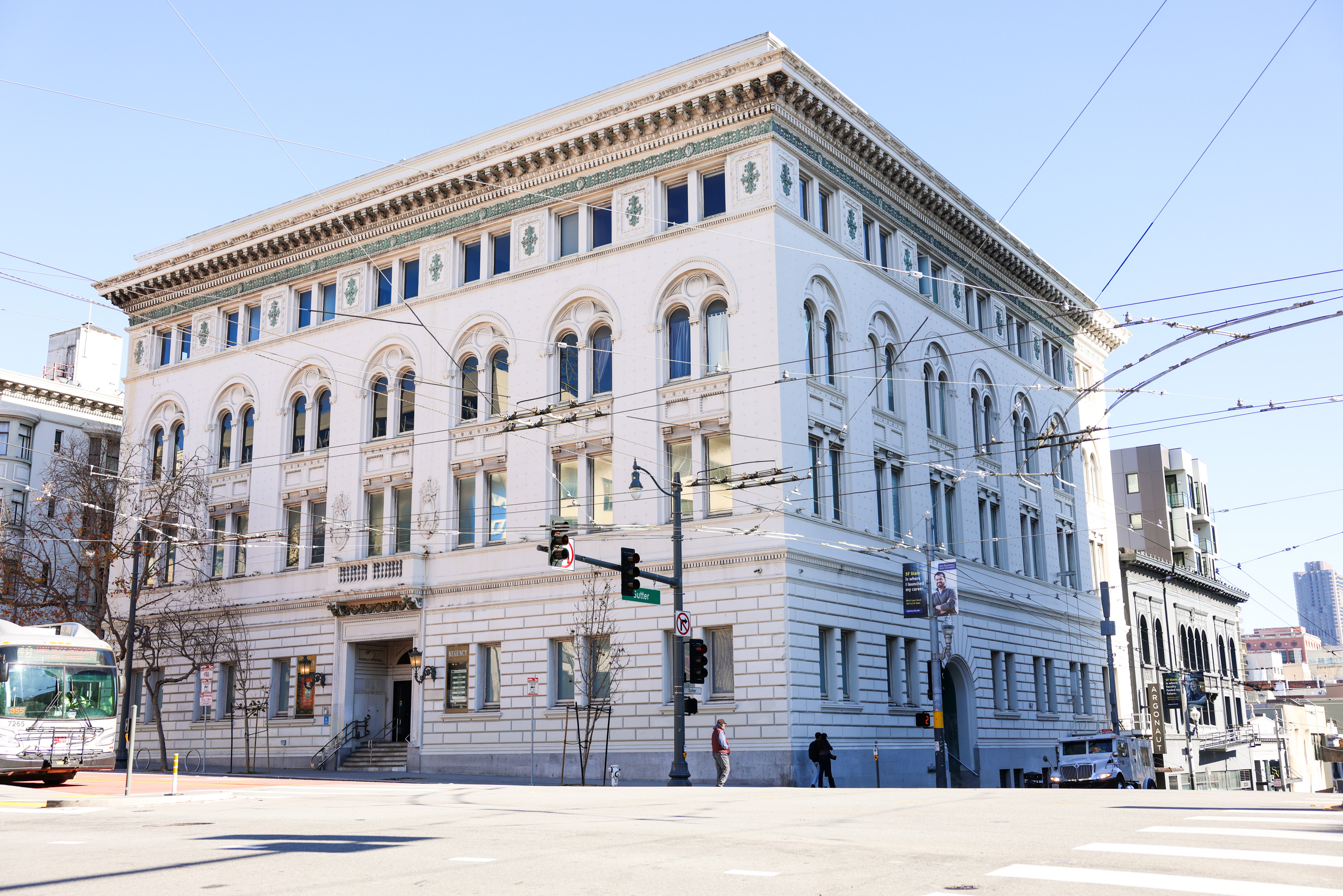 The image shows a large, ornate, corner building with arched windows and decorative stonework. It's located at a street intersection under a clear blue sky.