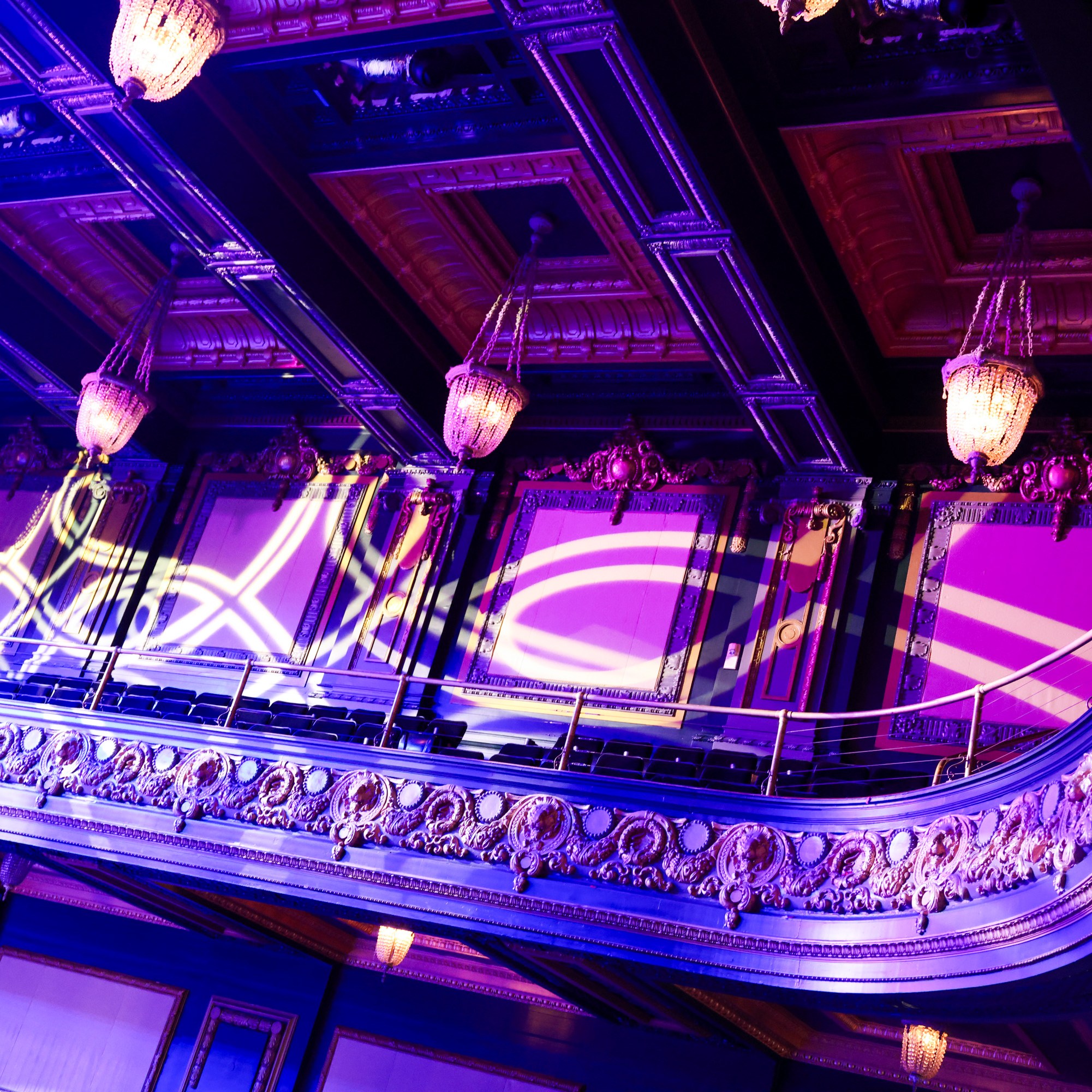 The image shows an ornate balcony in a theater with intricate moldings, chandeliers, and purple lighting. Decorative light patterns are projected on the walls.