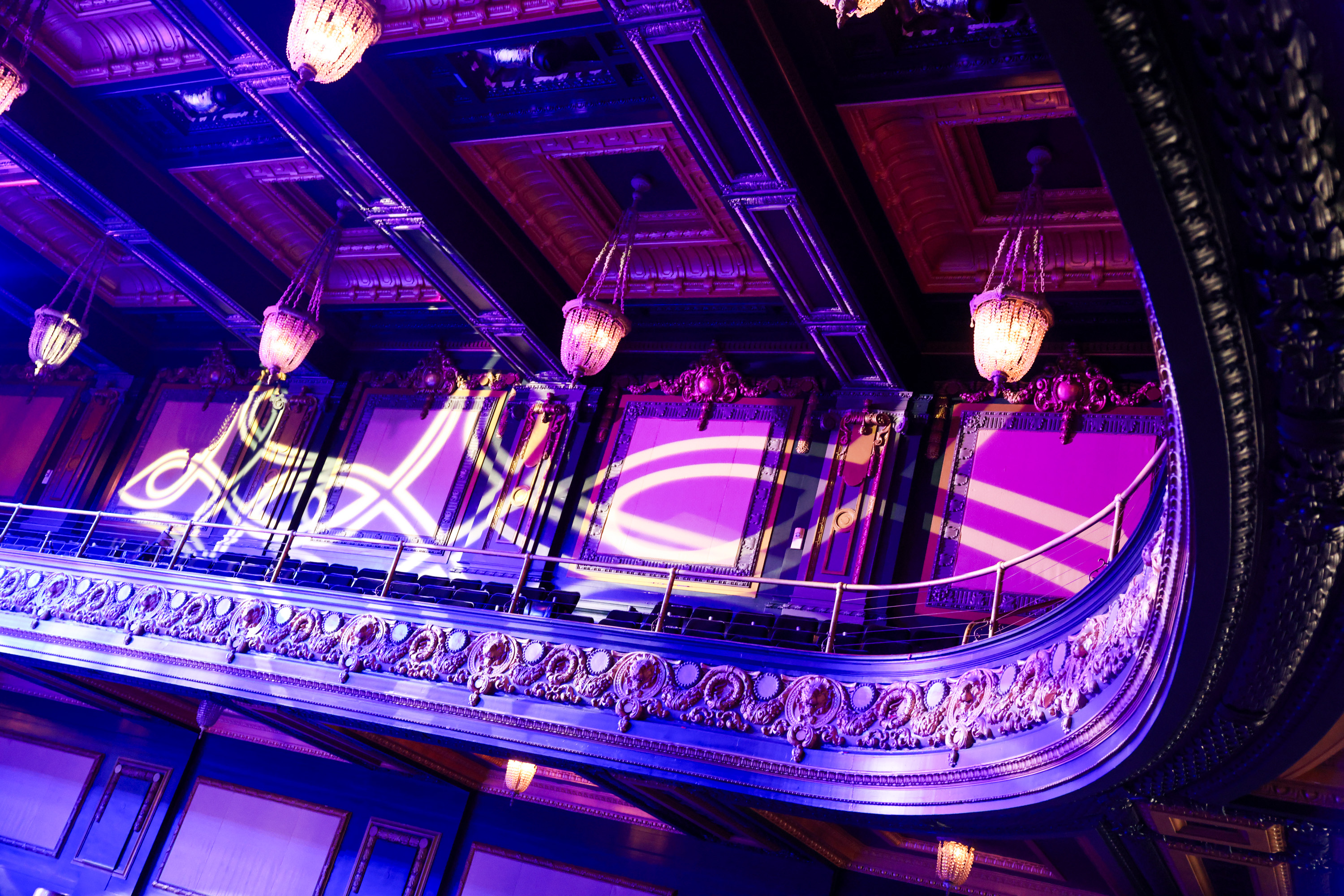 The image shows an ornate balcony in a theater with intricate moldings, chandeliers, and purple lighting. Decorative light patterns are projected on the walls.