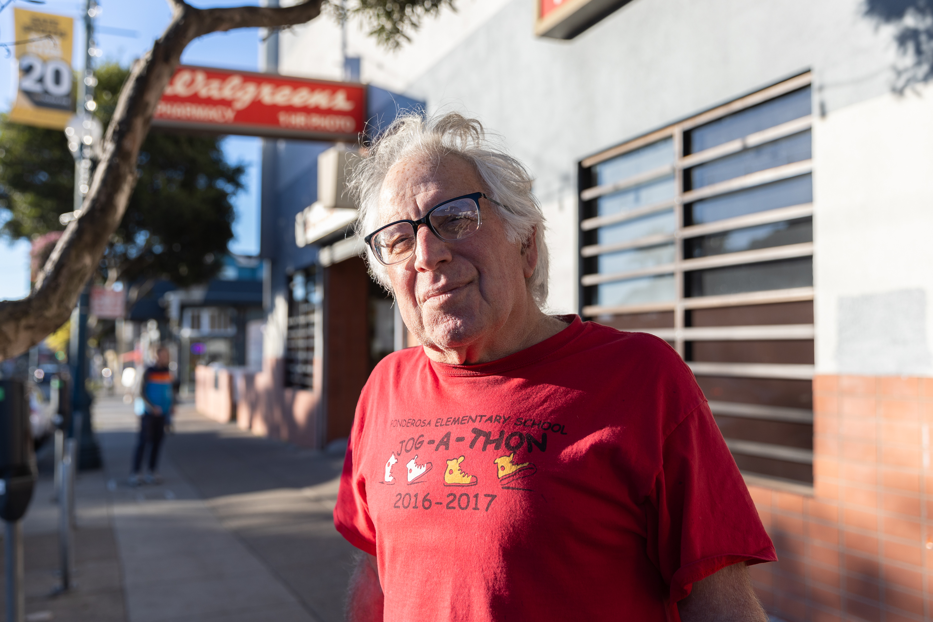 An older man with glasses and white hair stands on a sunny sidewalk, wearing a red T-shirt with &quot;Jog-a-thon 2016-2017&quot; printed on it.