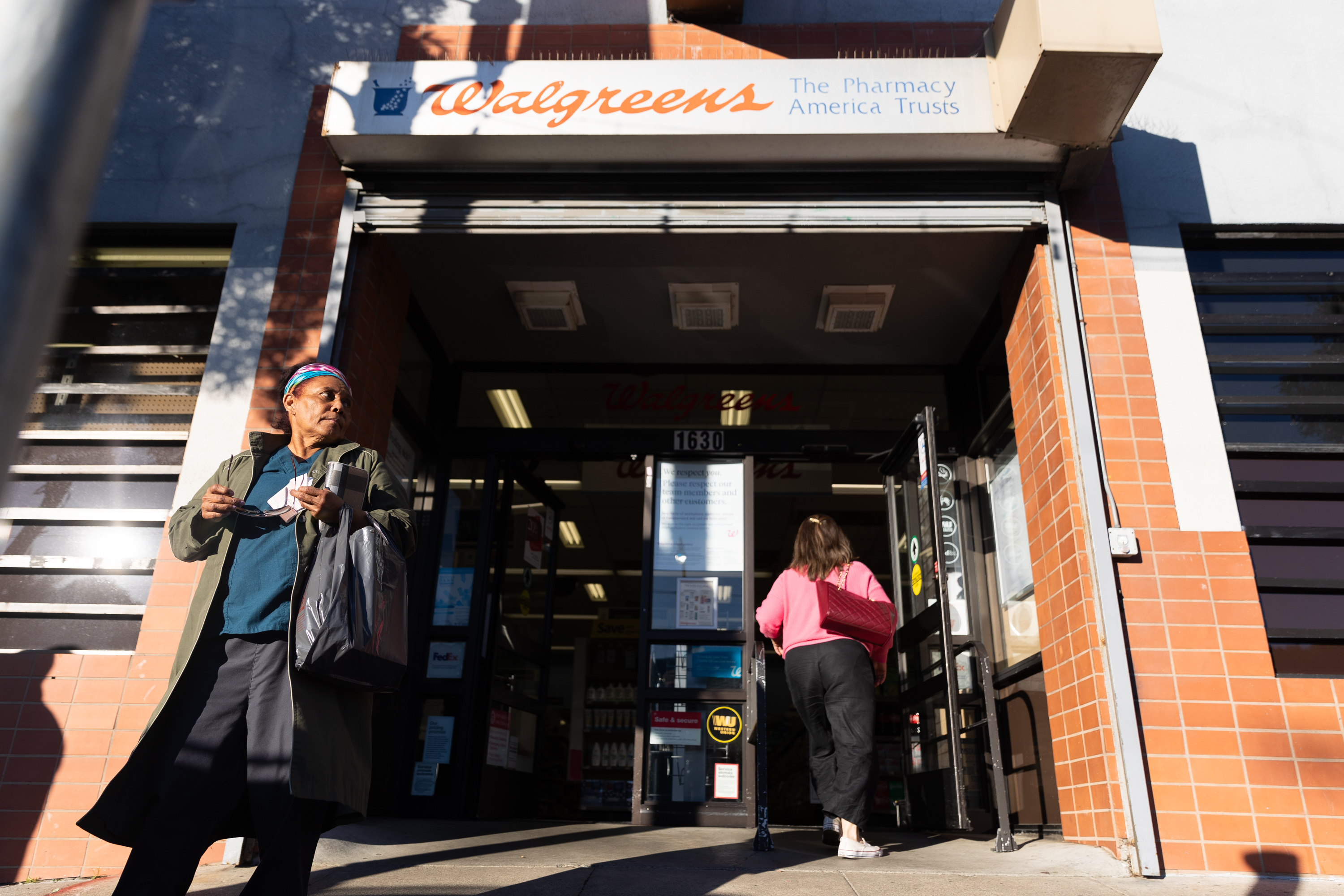 The image shows a Walgreens entrance with a person walking out holding a bag and another person entering the store. The building is brick with visible signage.