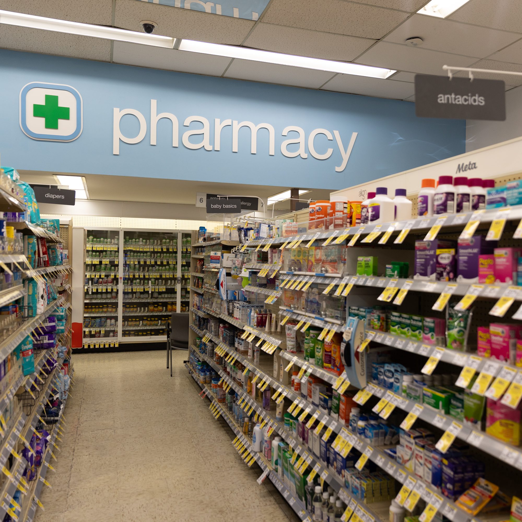 A pharmacy aisle filled with various health products, including antacids. Signs indicate sections for diapers and baby basics under bright overhead lighting.