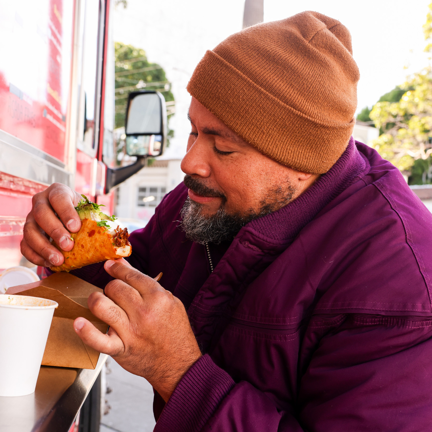 A man in a brown beanie and purple jacket enjoys a taco beside a red food truck, holding it close to his mouth while smiling. A drink is on the counter.