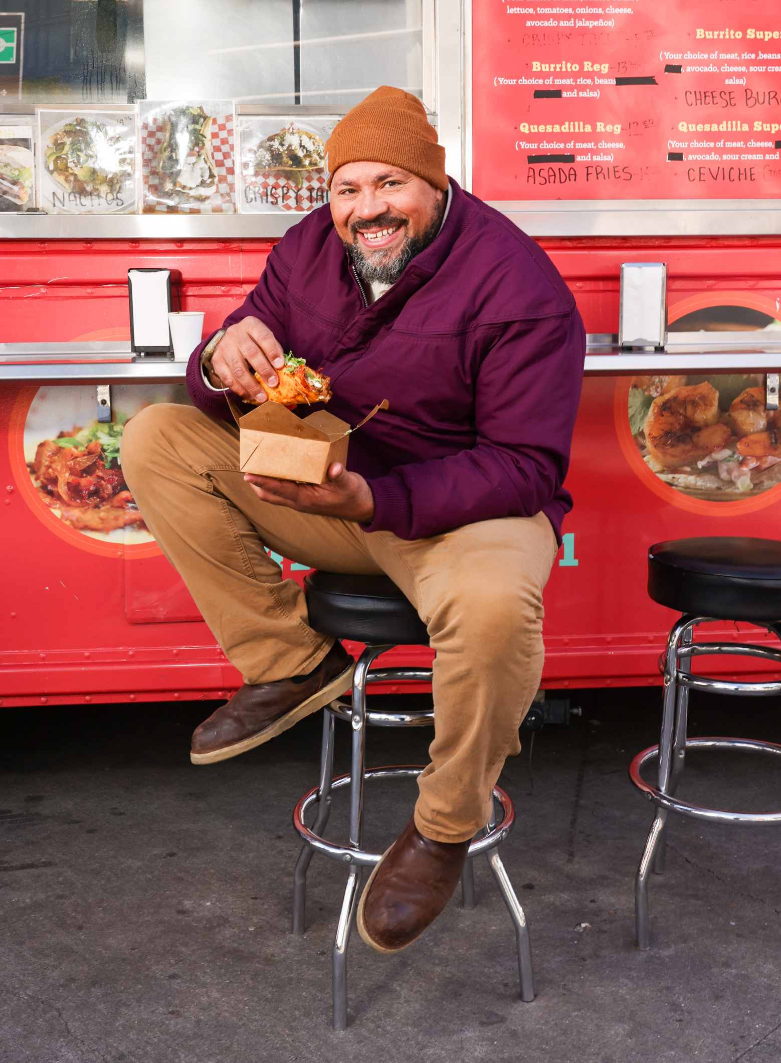 A man in a purple jacket and brown beanie joyfully holds a taco while sitting on a stool in front of a red food truck with menu items displayed.