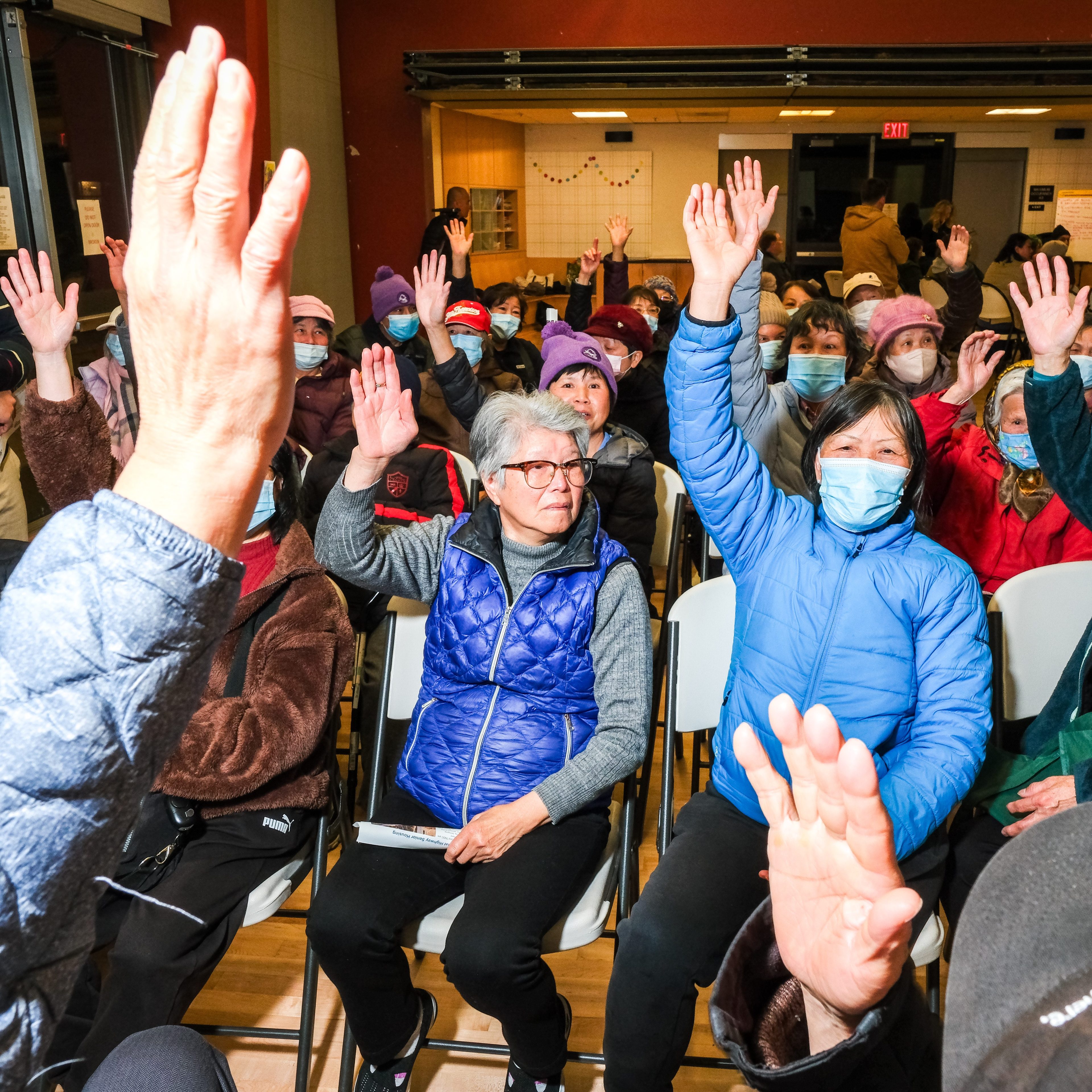 A group of people, mostly in winter coats and some with masks, are seated and raising their hands in a room, possibly during a meeting or event.