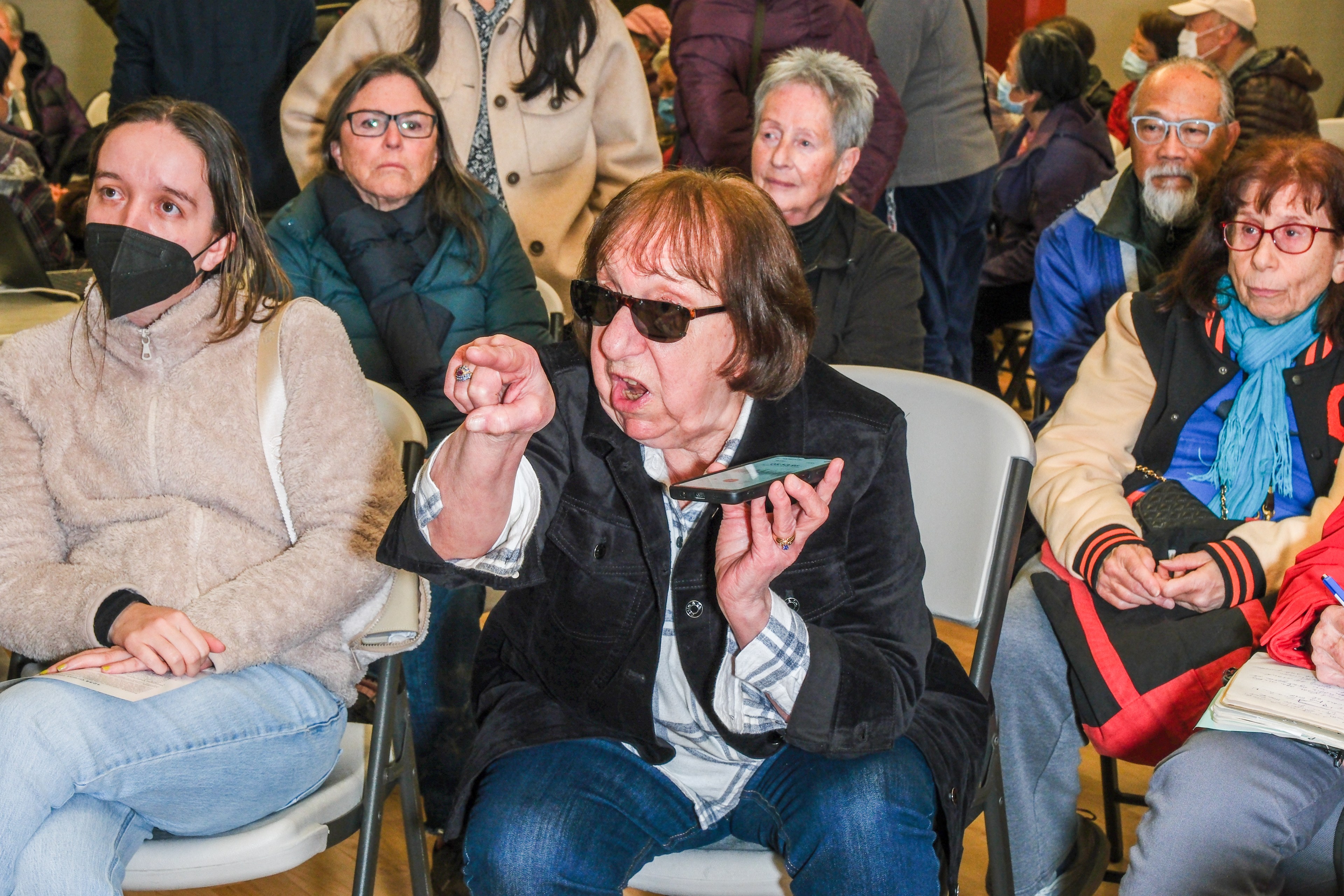 A group of people is seated indoors, with a woman in sunglasses animatedly pointing and holding a phone. Others appear attentive, and one wears a mask.