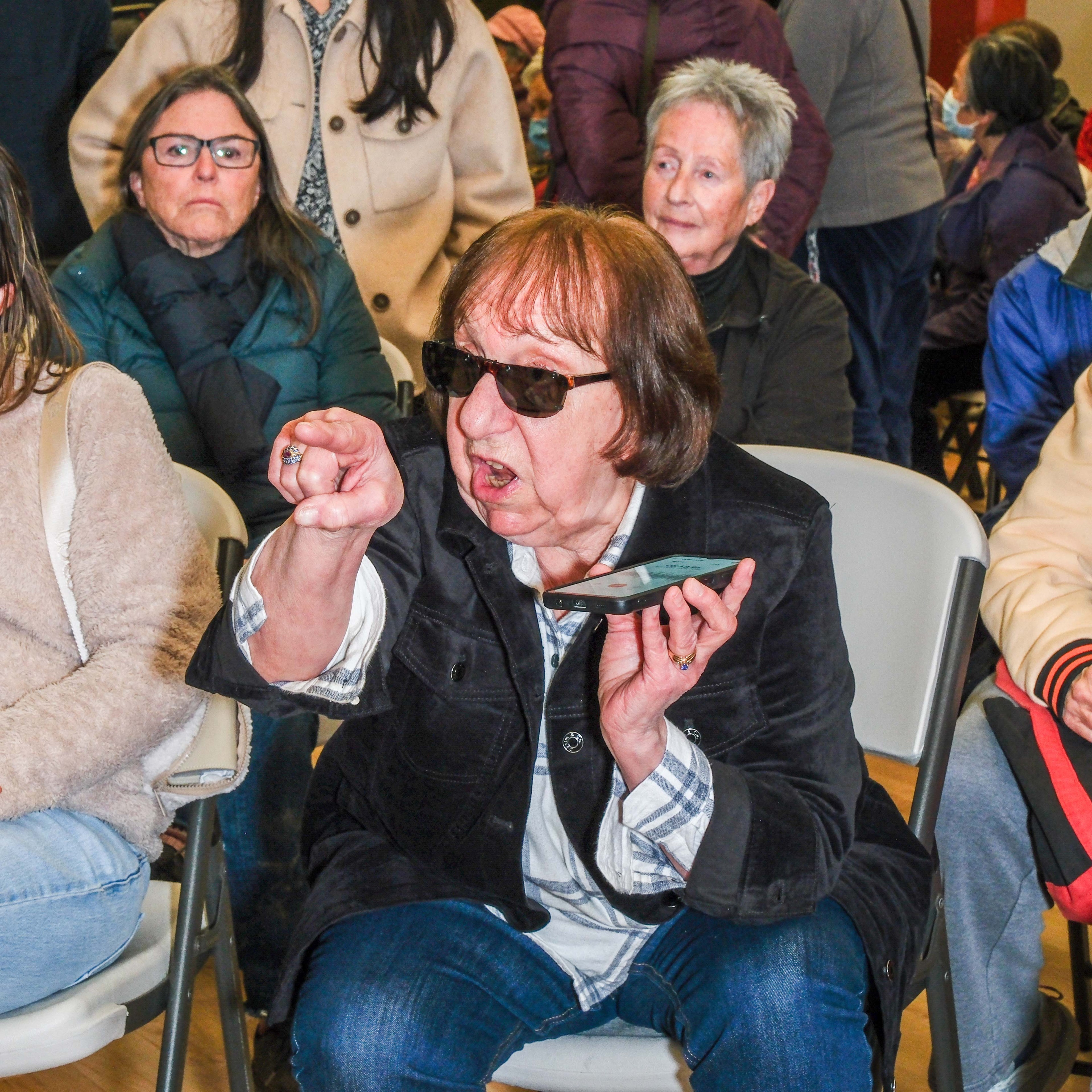 A woman wearing sunglasses sits on a chair, pointing her finger and looking upset. She holds a phone in her other hand. Behind her, other seated people watch.