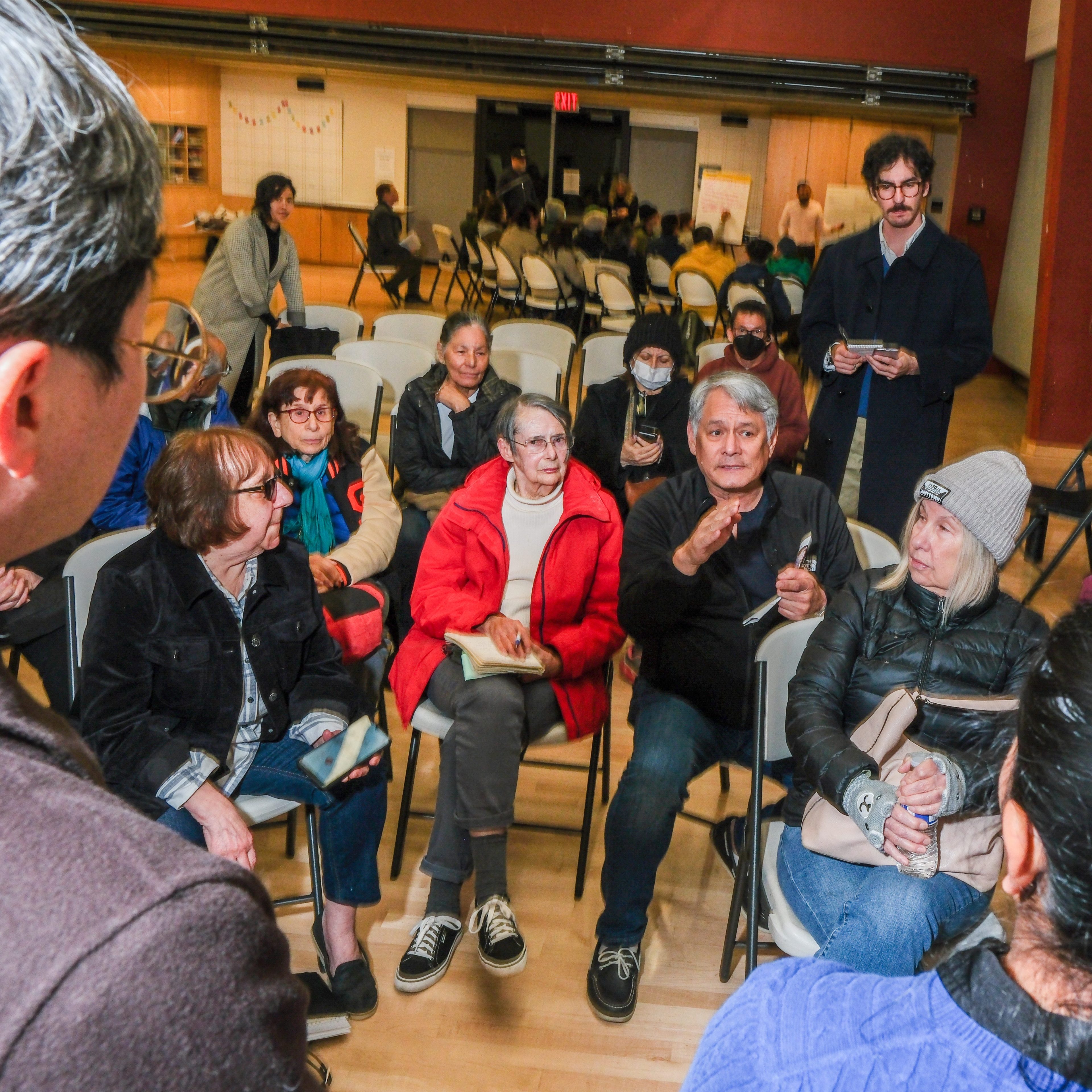 A diverse group of people sits in a circular formation, engaging in a discussion. Some listen attentively, while others take notes. The setting appears to be a community hall.