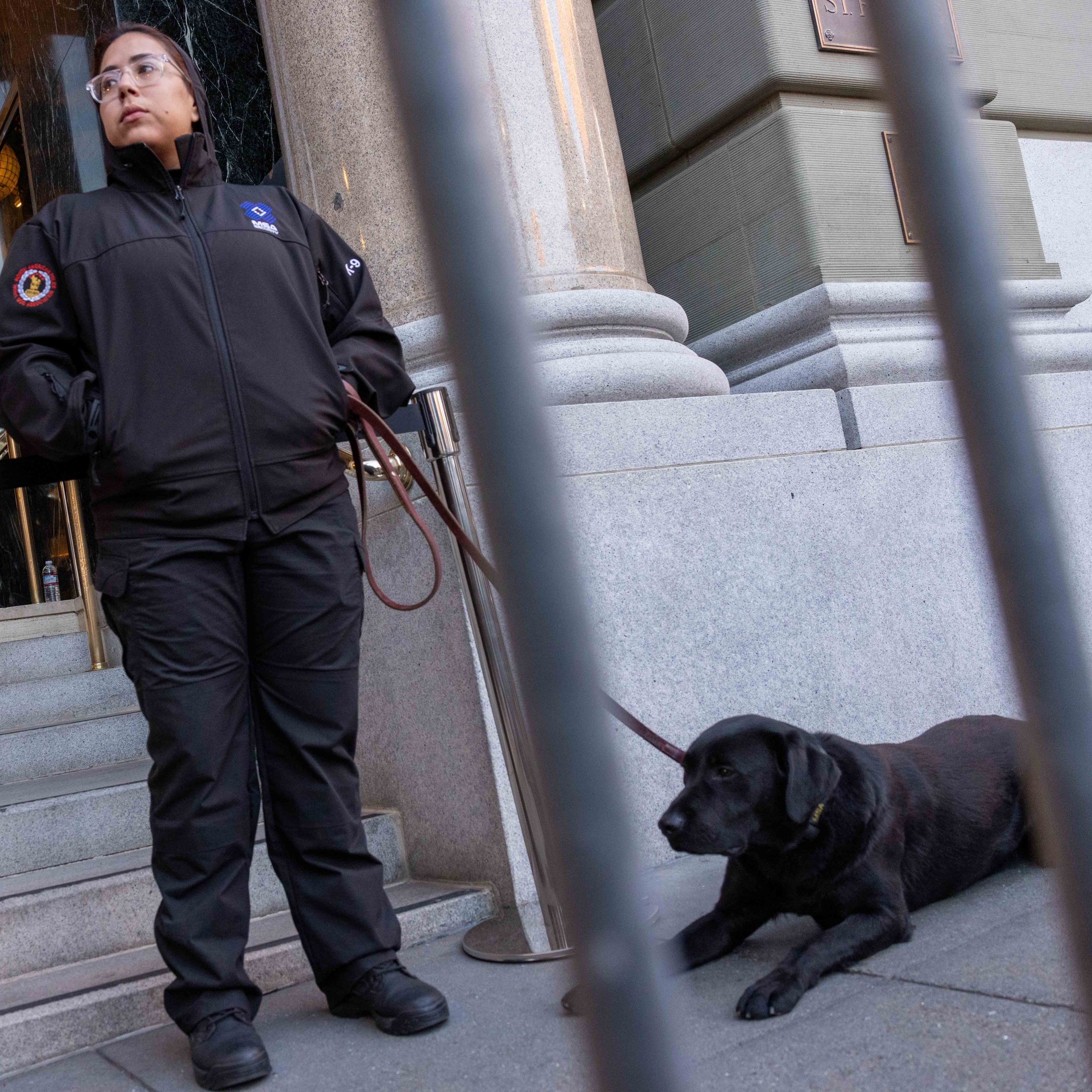 A security officer stands near a building entrance, holding a leash attached to a black dog lying on the ground. Another officer stands on nearby steps.