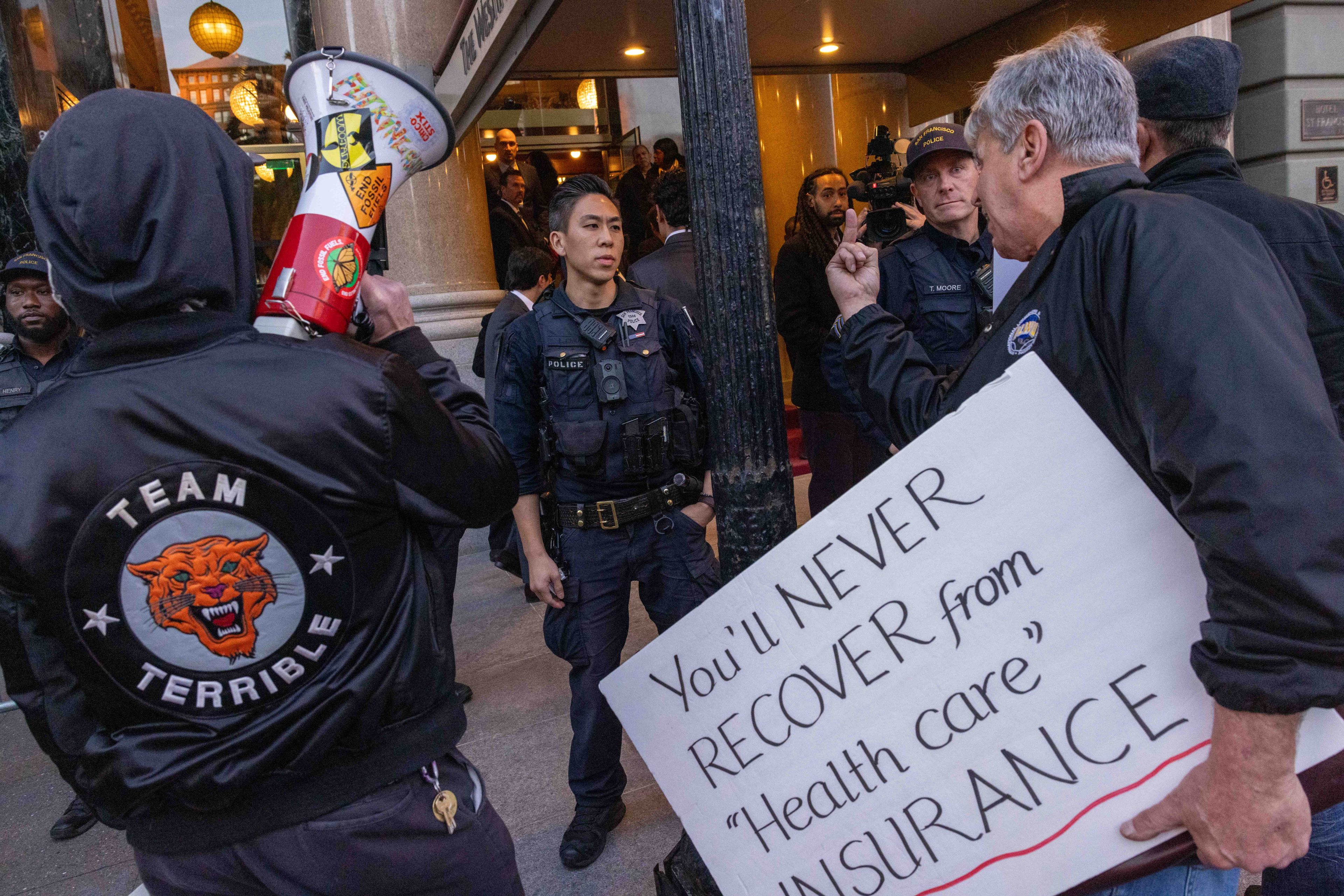 Protesters, including one with a megaphone and jacket saying &quot;Team Terrible,&quot; hold a sign reading &quot;You'll NEVER RECOVER from 'Health care' INSURANCE&quot; facing police.