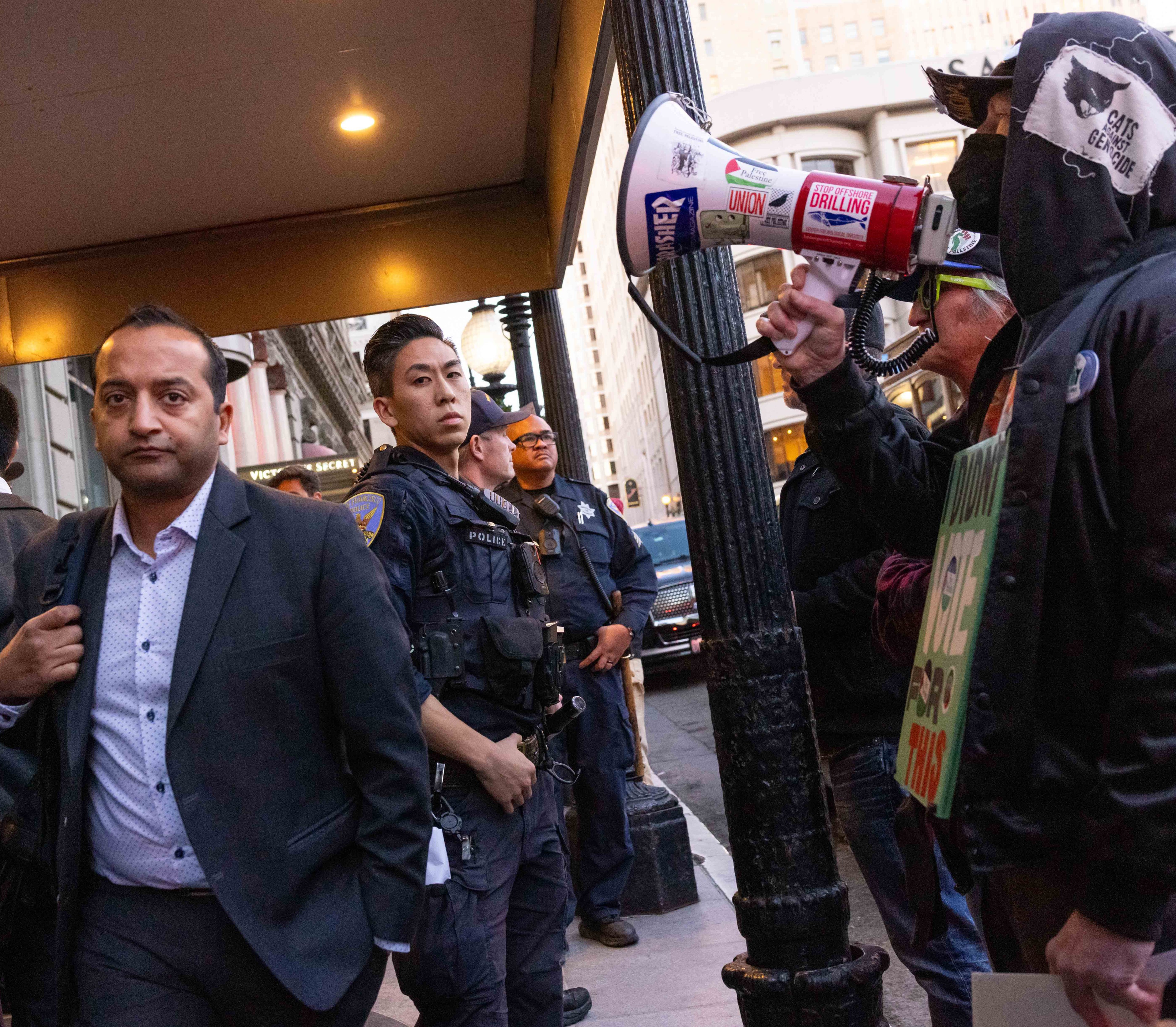 A man walks past police officers on a city street. Nearby, a person holds a megaphone covered in protest stickers, speaking to a small crowd.