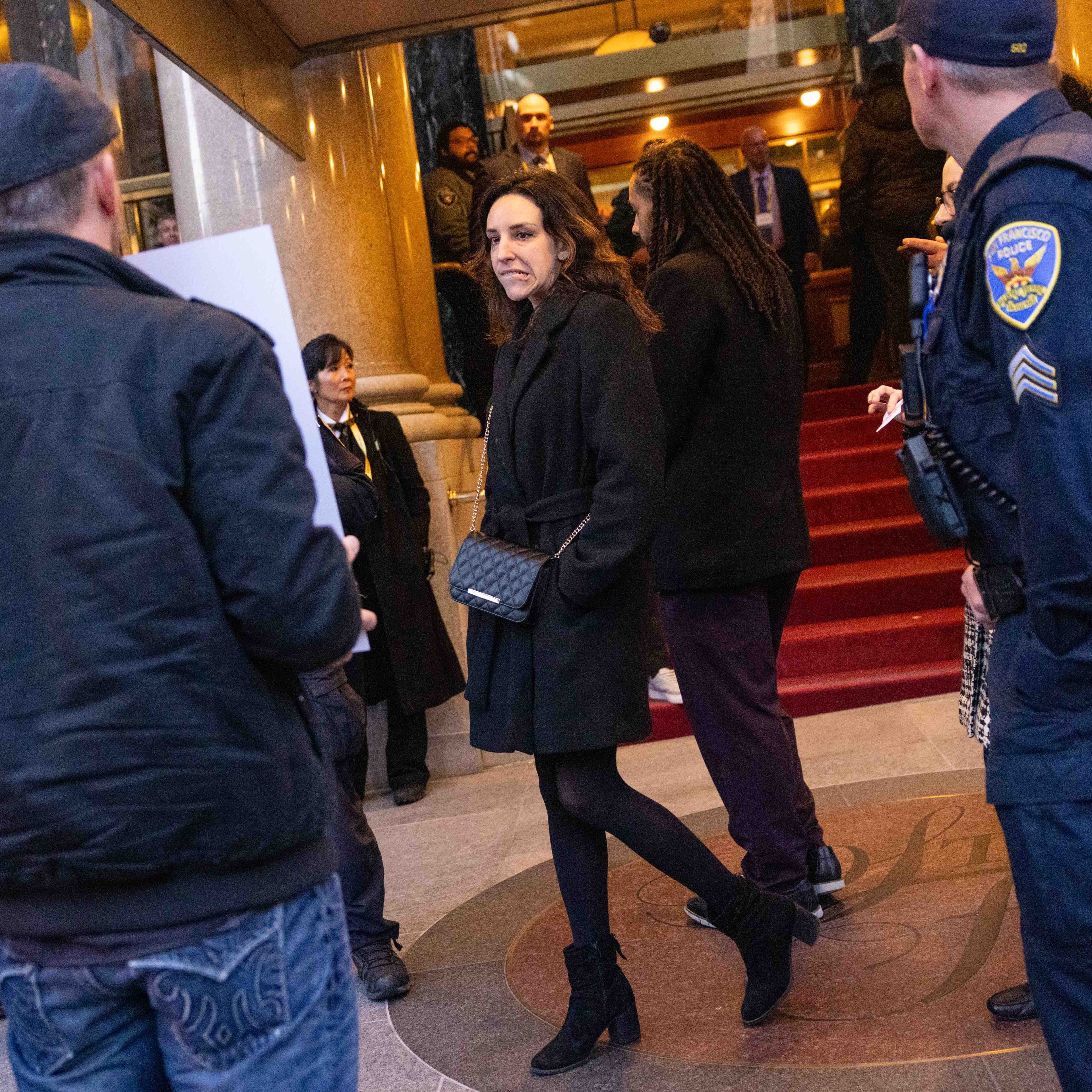 A woman in a black coat stands by red carpeted steps, surrounded by people, including a police officer. The scene appears to be outside a building entrance.