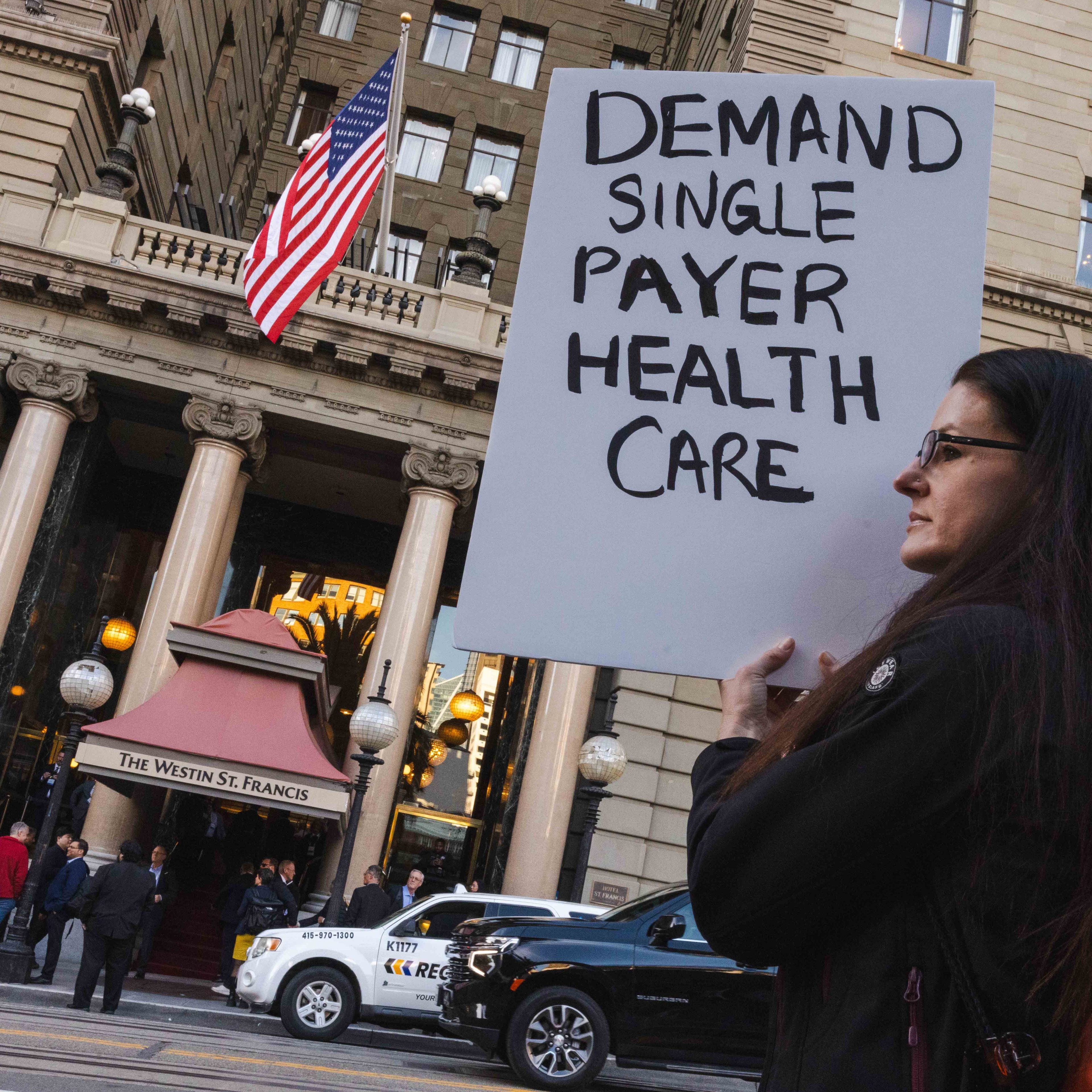 A person holds a sign reading &quot;DEMAND SINGLE PAYER HEALTH CARE&quot; outside a building with columns and an American flag. Several people and cars are nearby.