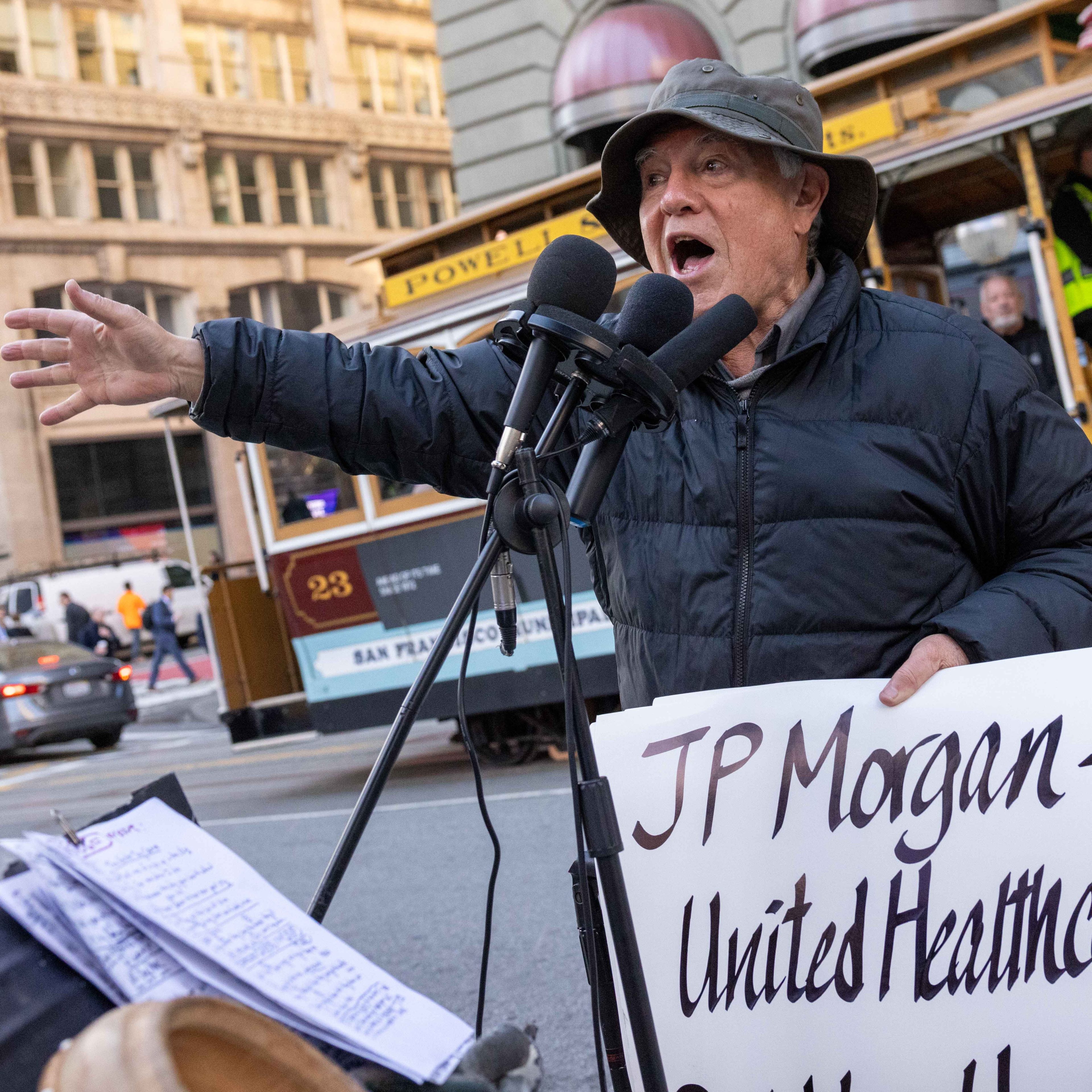 A man in a hat and jacket speaks passionately into microphones, holding a protest sign, with a San Francisco cable car and city buildings in the background.