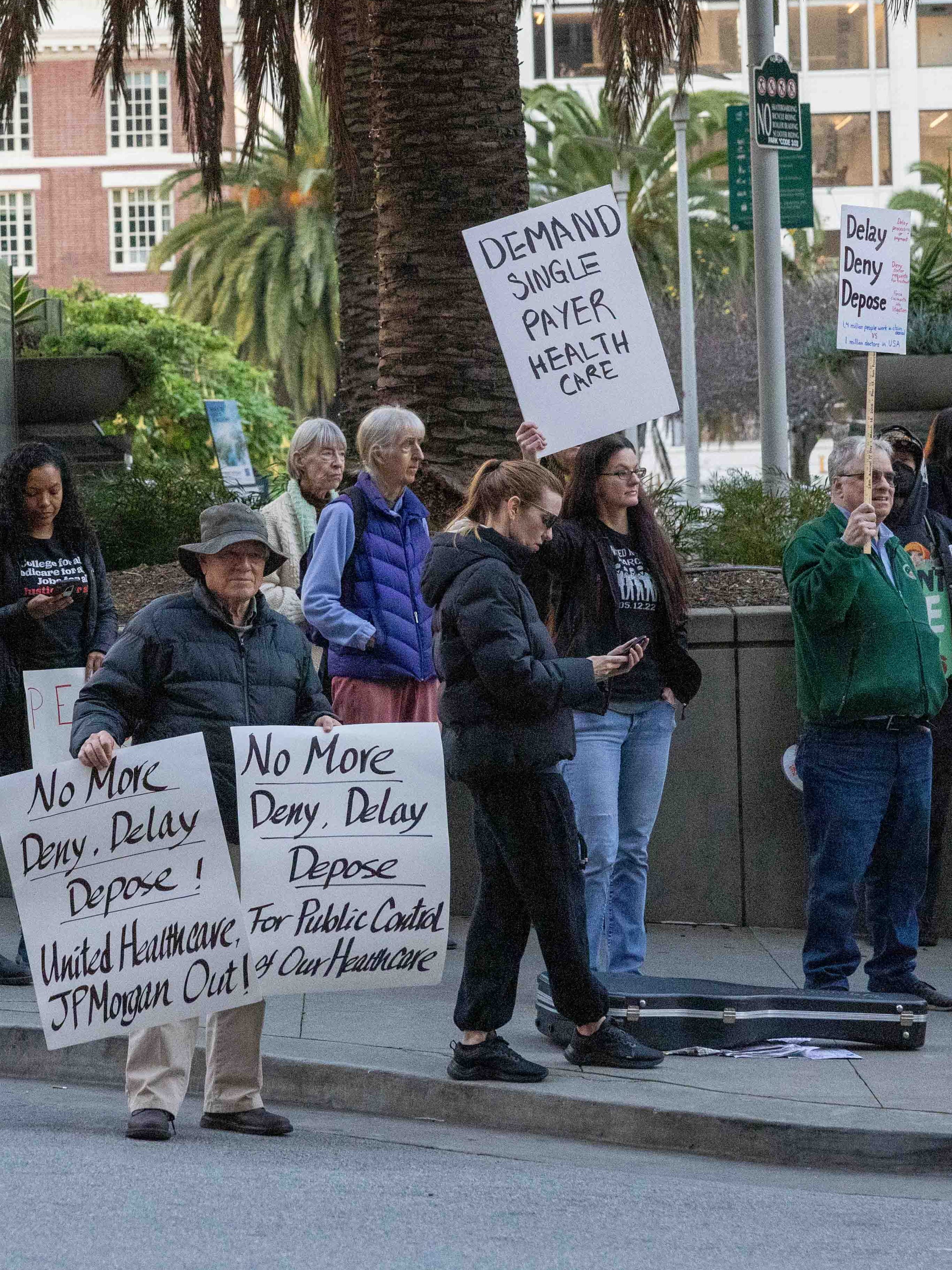 A group of people protest on a city sidewalk, holding signs advocating for healthcare reform. Palms and buildings serve as the urban backdrop.