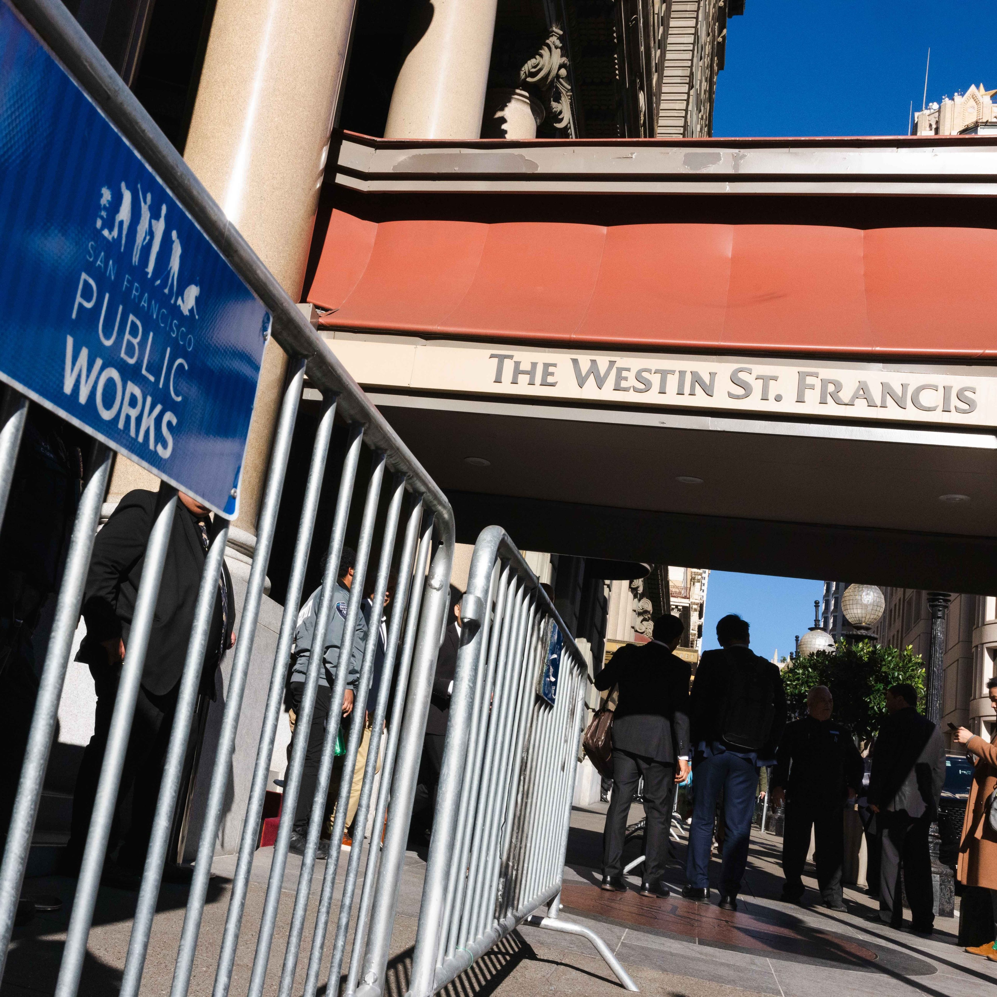 Metal barricades with a &quot;San Francisco Public Works&quot; sign are set up outside The Westin St. Francis hotel entrance, with several people walking nearby.