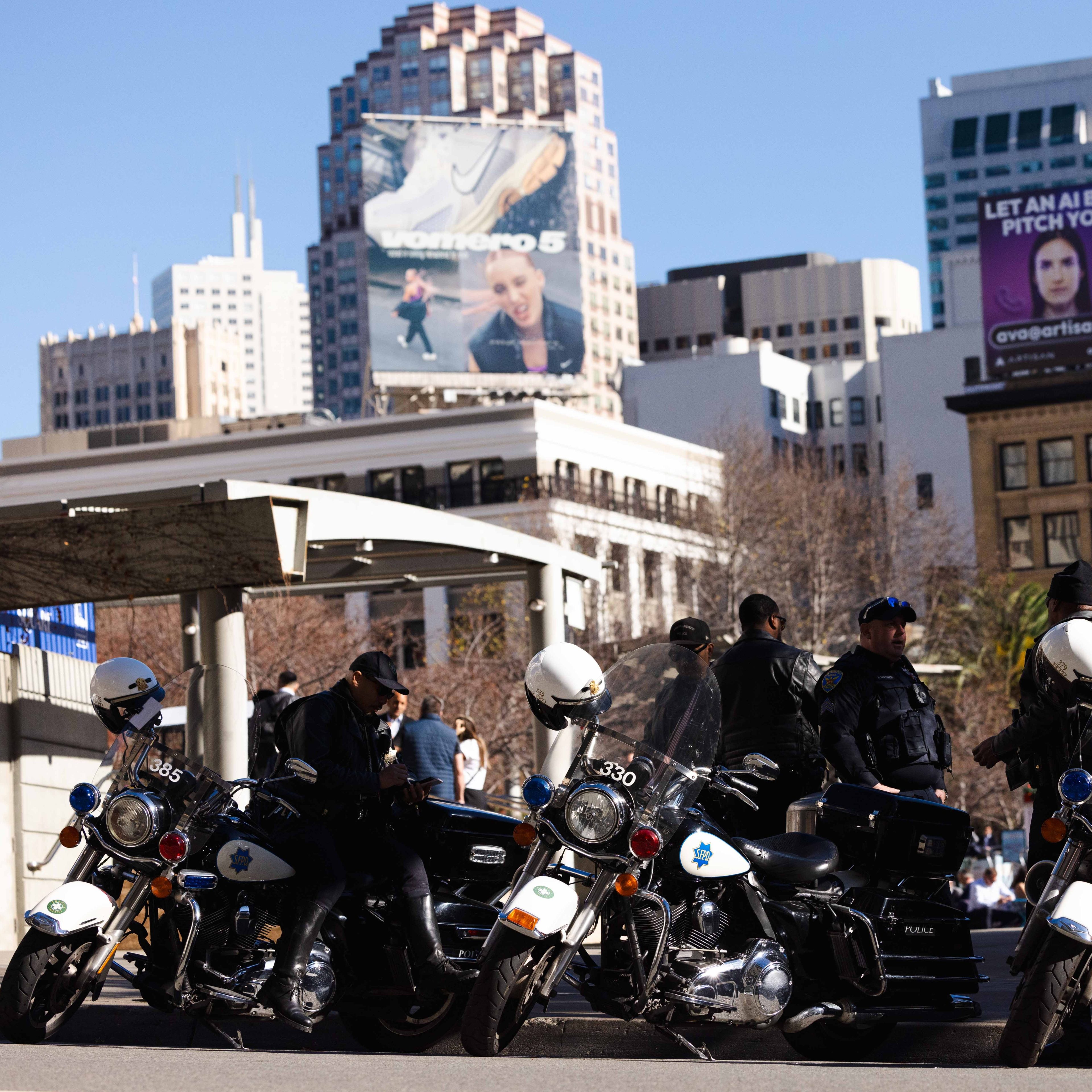 Several police officers and motorcycles are in the foreground, with a cityscape and billboards in the background under a clear blue sky.