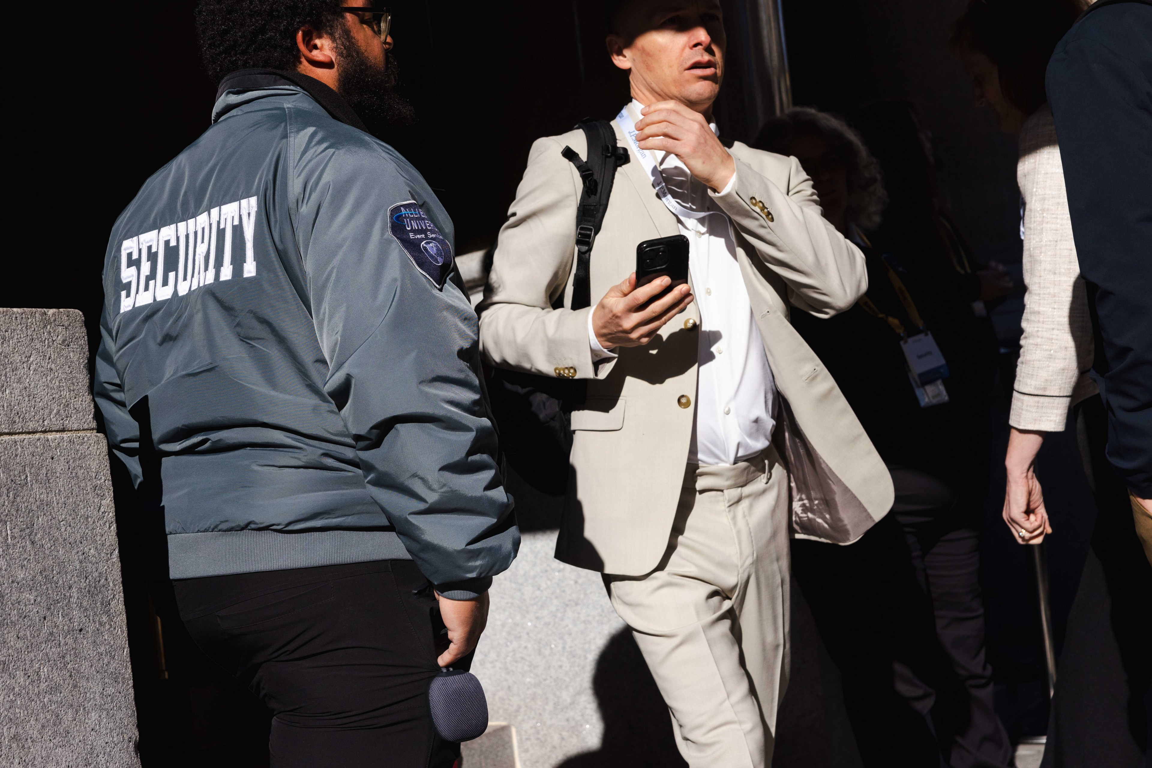 A man in a light suit holds a phone and walks briskly past a security guard in a blue jacket labeled "SECURITY." The scene is bustling with several people.