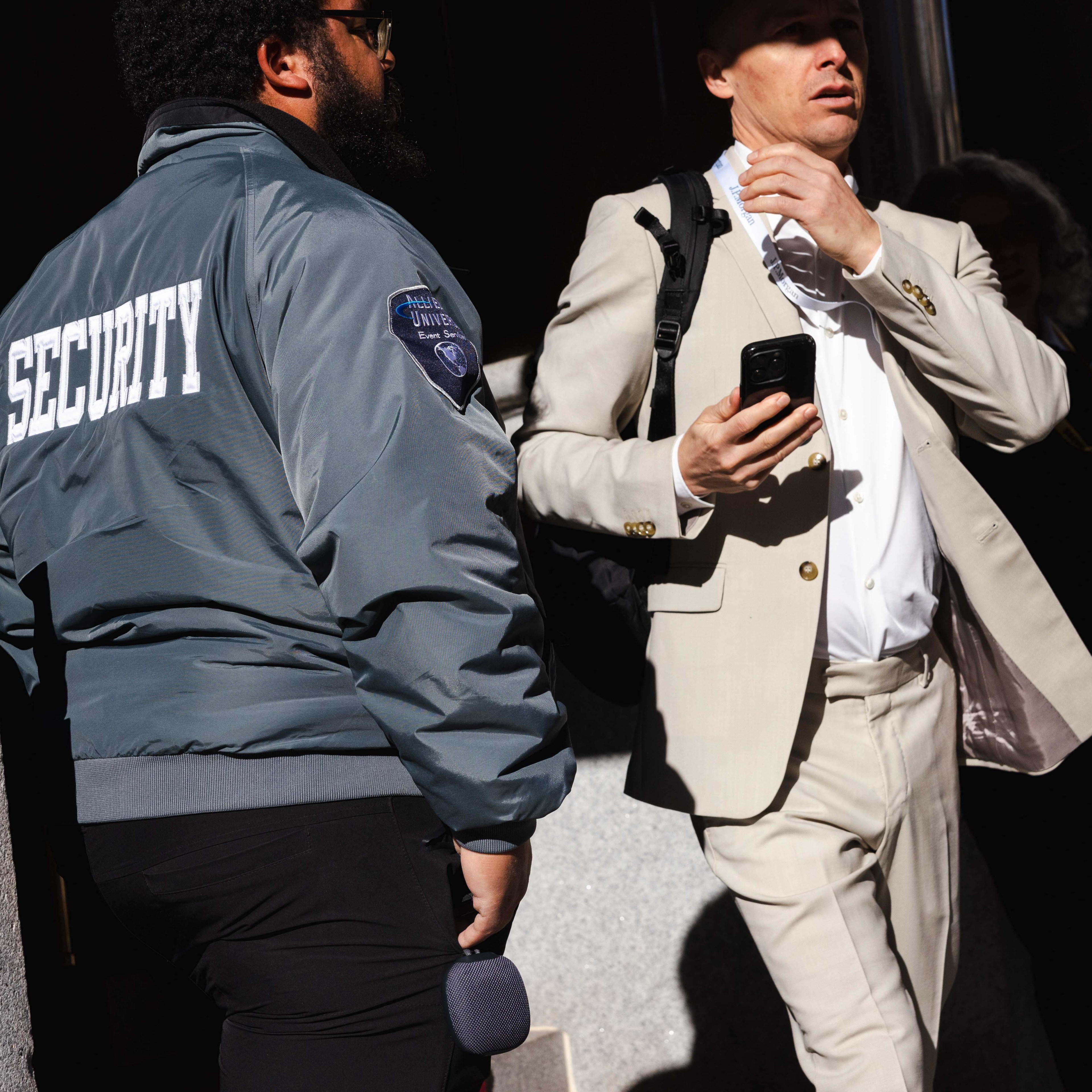 A security guard wearing a blue jacket stands next to a man in a beige suit holding a phone and adjusting his tie. They are outside in bright sunlight.