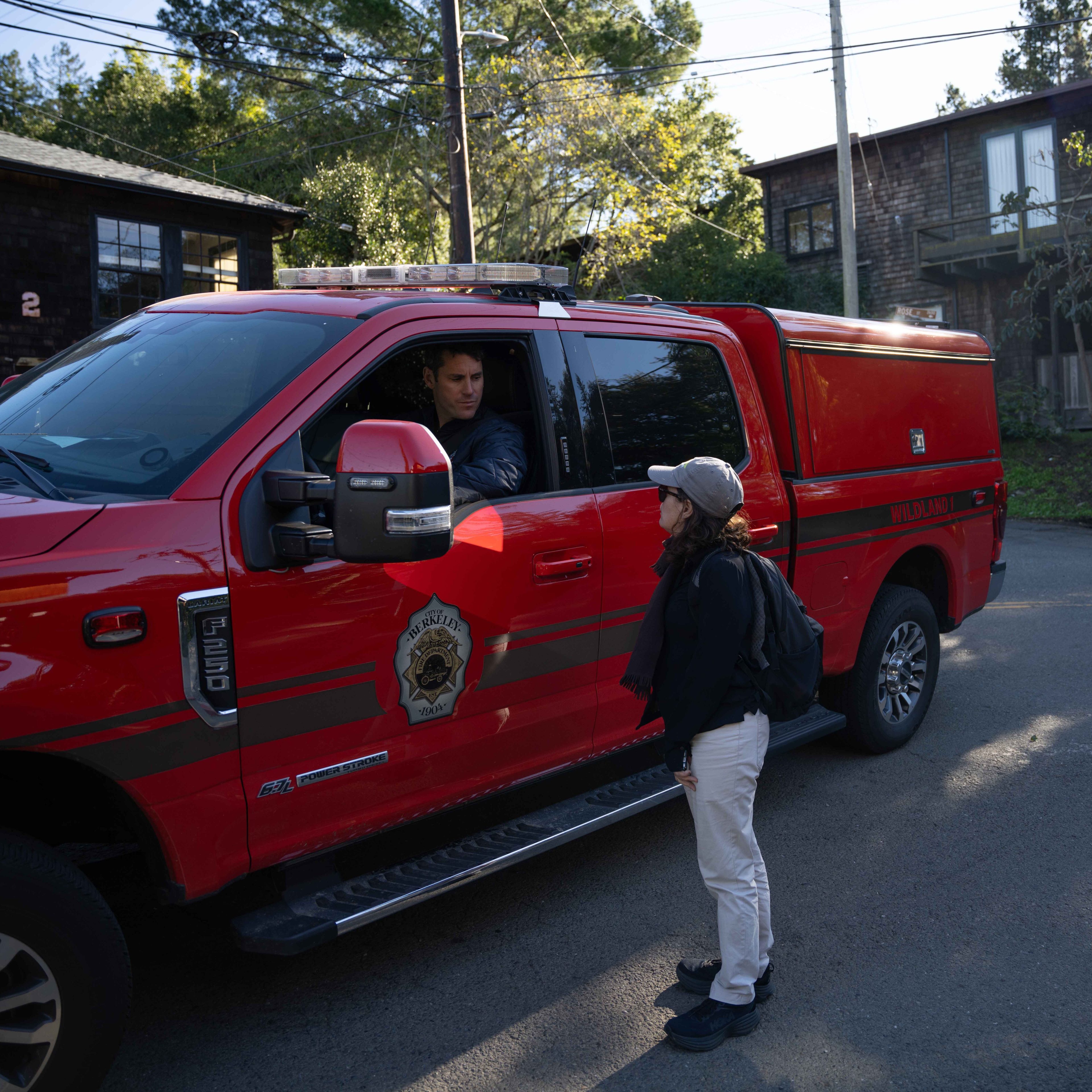 A woman stands talking to a person in a red emergency vehicle labeled &quot;Berkeley Fire.&quot; The vehicle is parked on a residential street with trees and houses nearby.