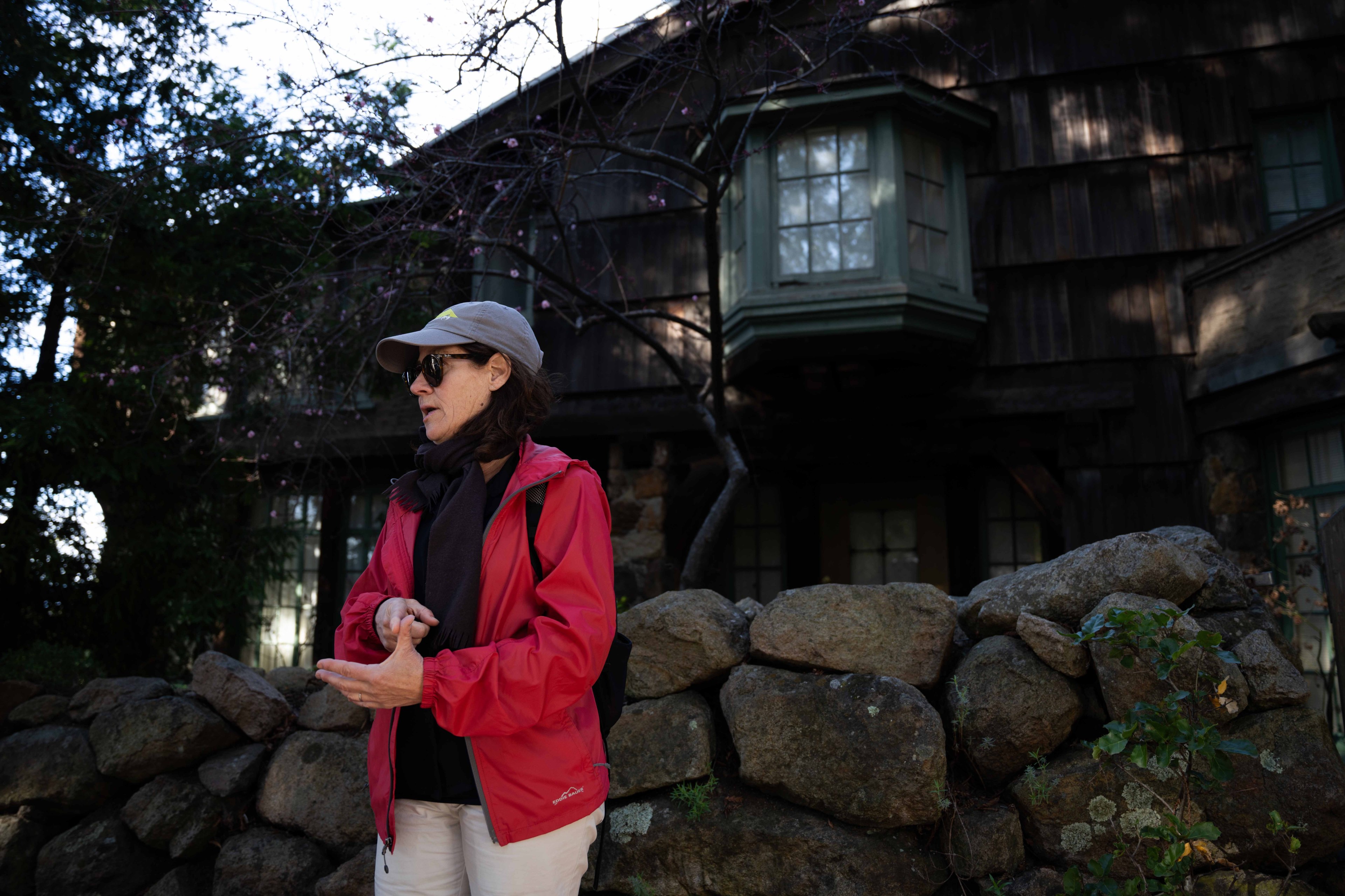 A person in a red jacket and cap stands near a stone wall, gesturing with their hands. Behind them is a rustic building with a large bay window.