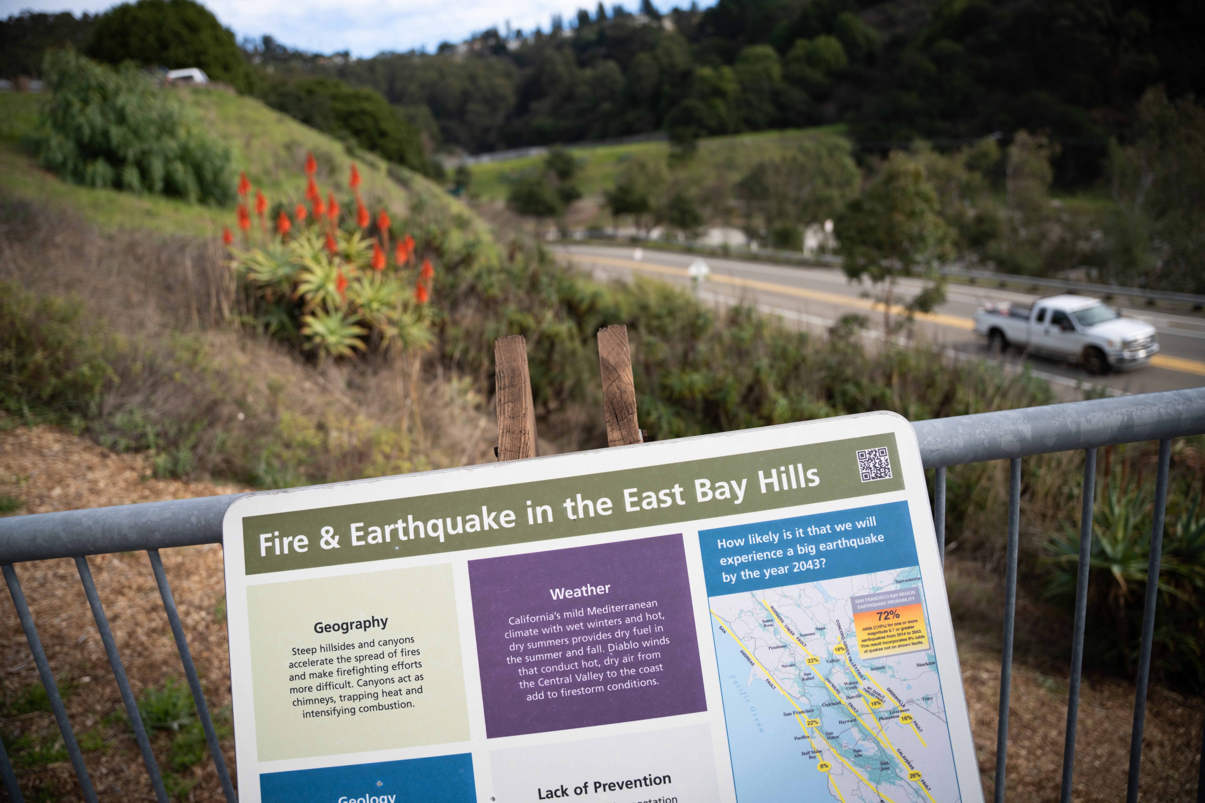 A sign titled &quot;Fire &amp; Earthquake in the East Bay Hills&quot; stands near a roadside with hilly greenery and a white truck passing on the road. Bright red flowers are visible nearby.