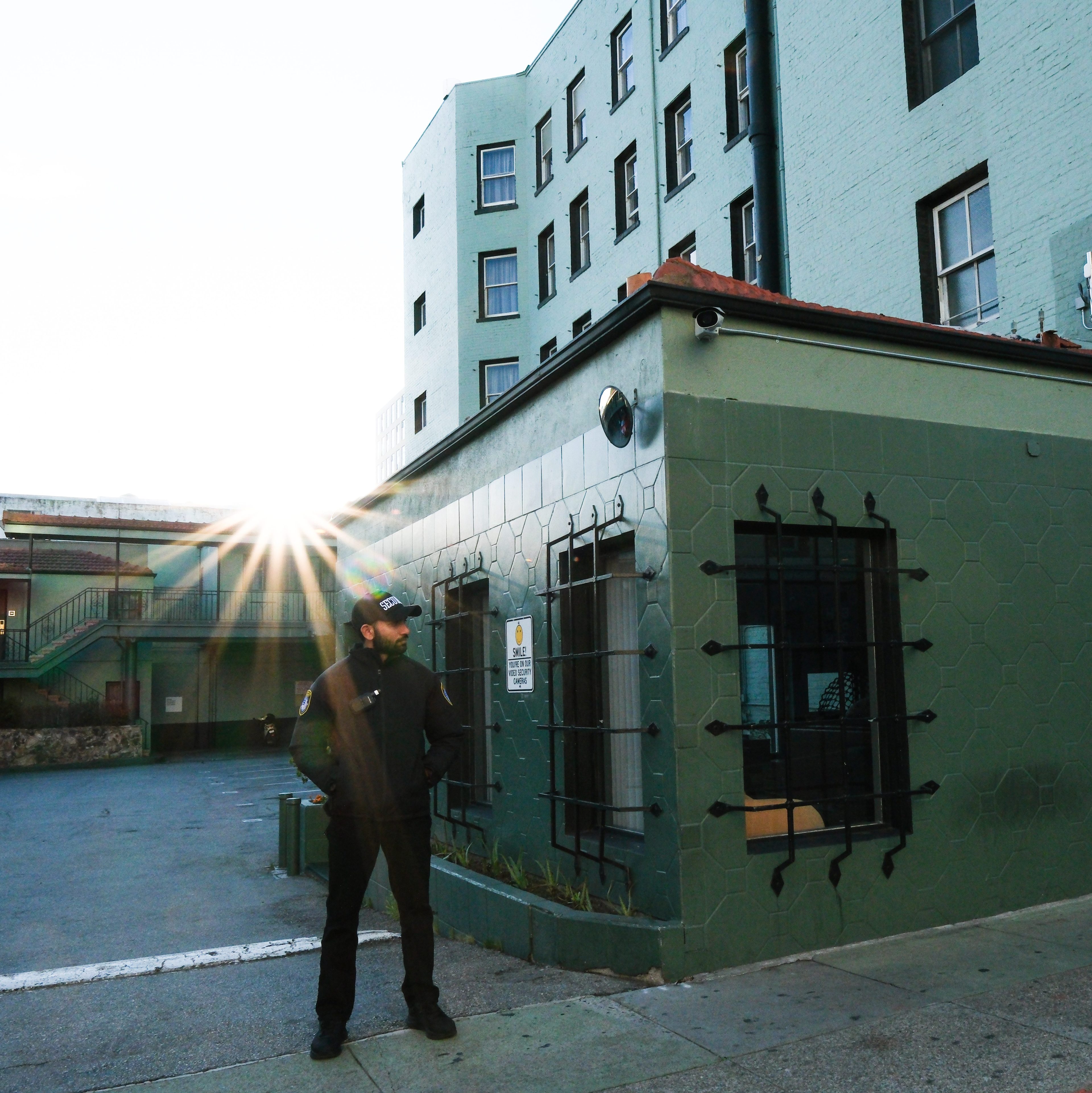 A person stands outside a green building with barred windows. Sunlight peeks behind them, creating a starburst effect. A multi-story building is in the background.