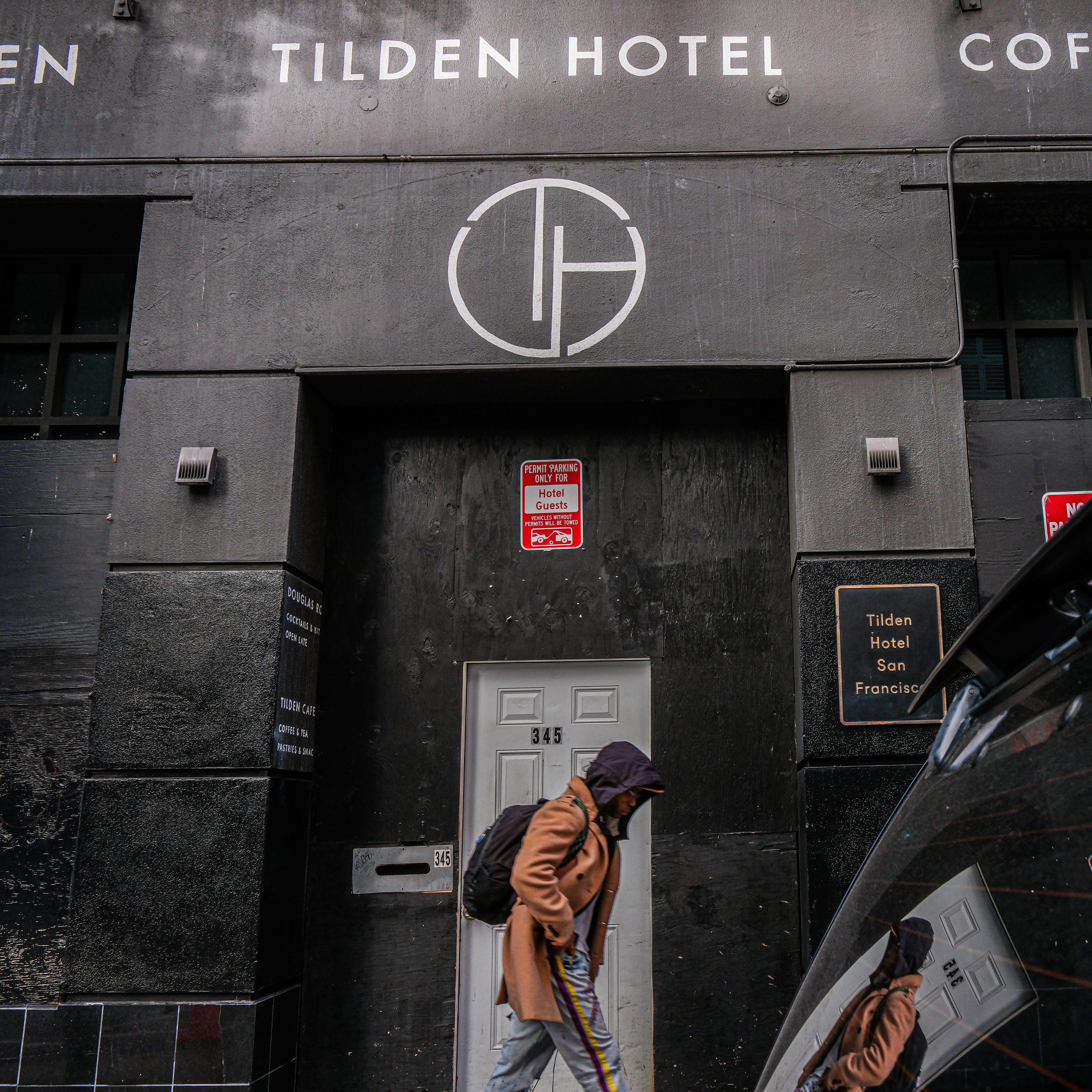 A person in a coat and hood walks past the entrance of Tilden Hotel. The building's facade is dark with signage and a distinct round logo above the door.