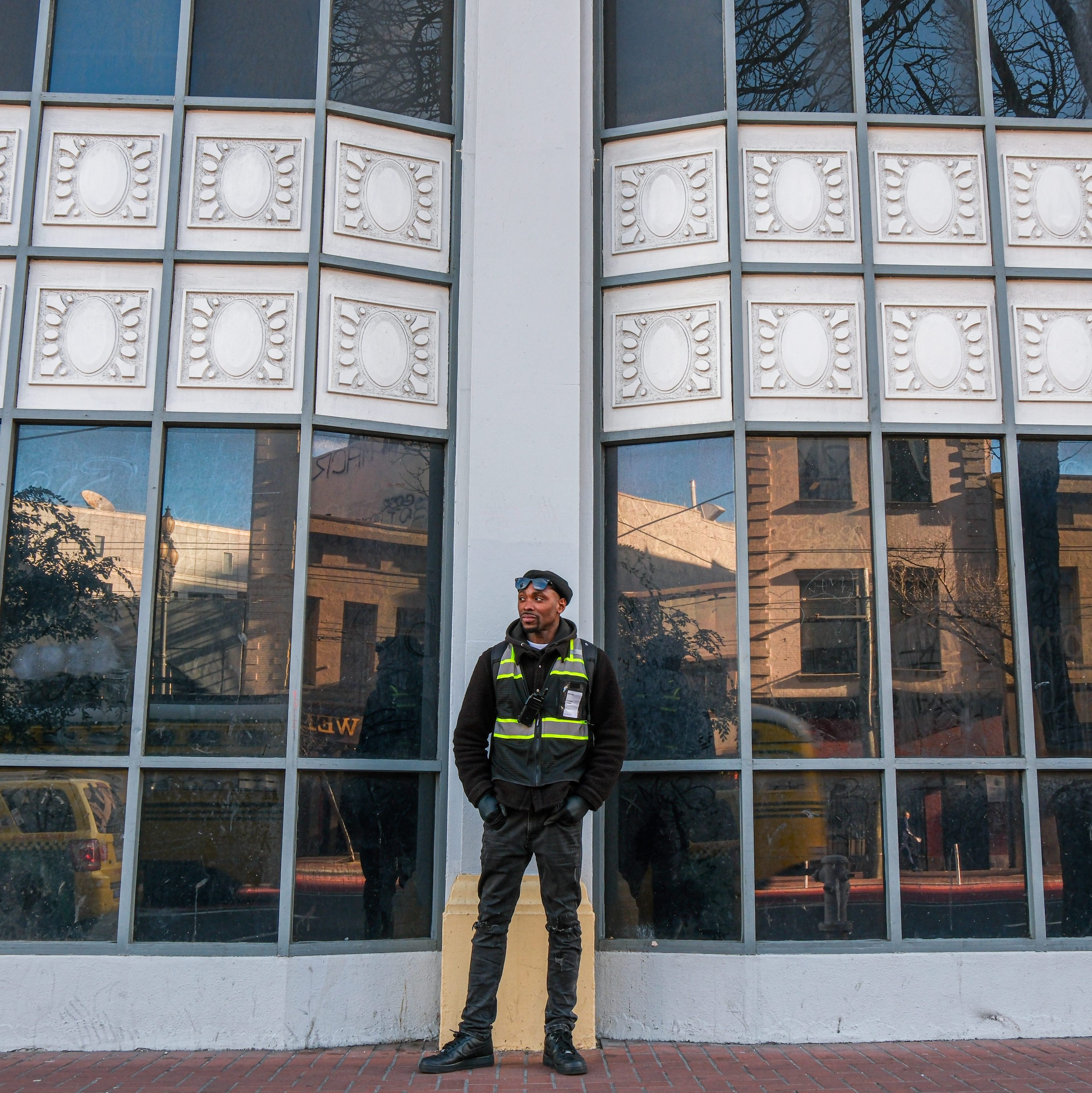 A person stands in front of tall, reflective windows, wearing a dark uniform and a high-visibility vest. The background shows a building facade and tree reflections.