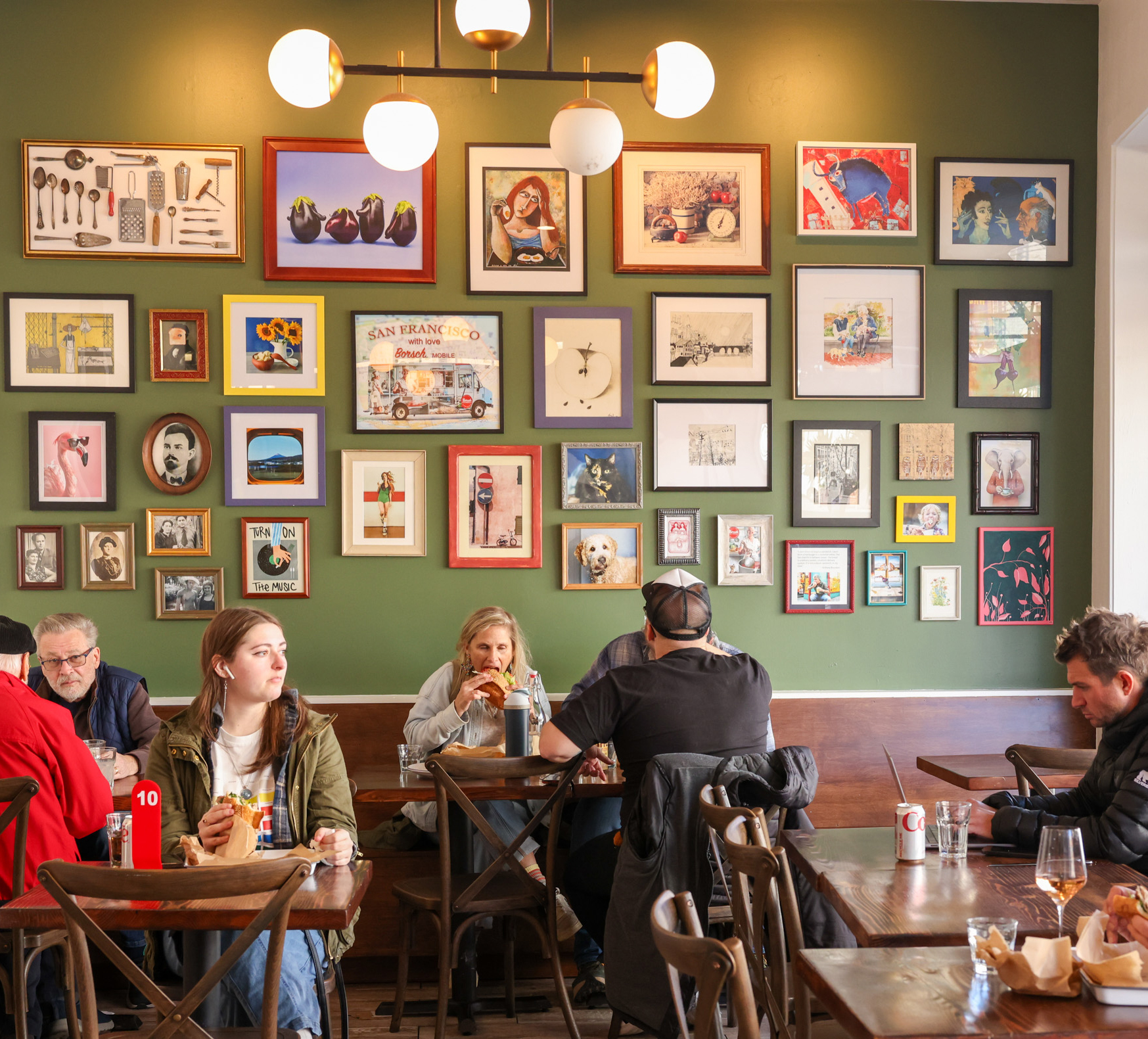 A cozy cafe with people eating and chatting. A green wall behind them is adorned with a diverse collection of framed artwork and photos, adding a vibrant touch.