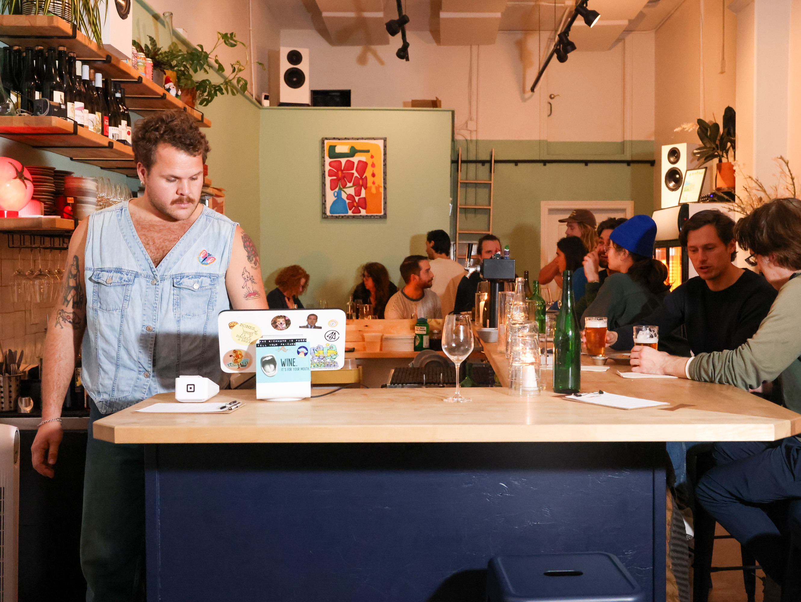 A bustling bar scene with a bartender in a denim vest working at a counter. Patrons are socializing and drinking at the bar. The atmosphere is lively and warm.