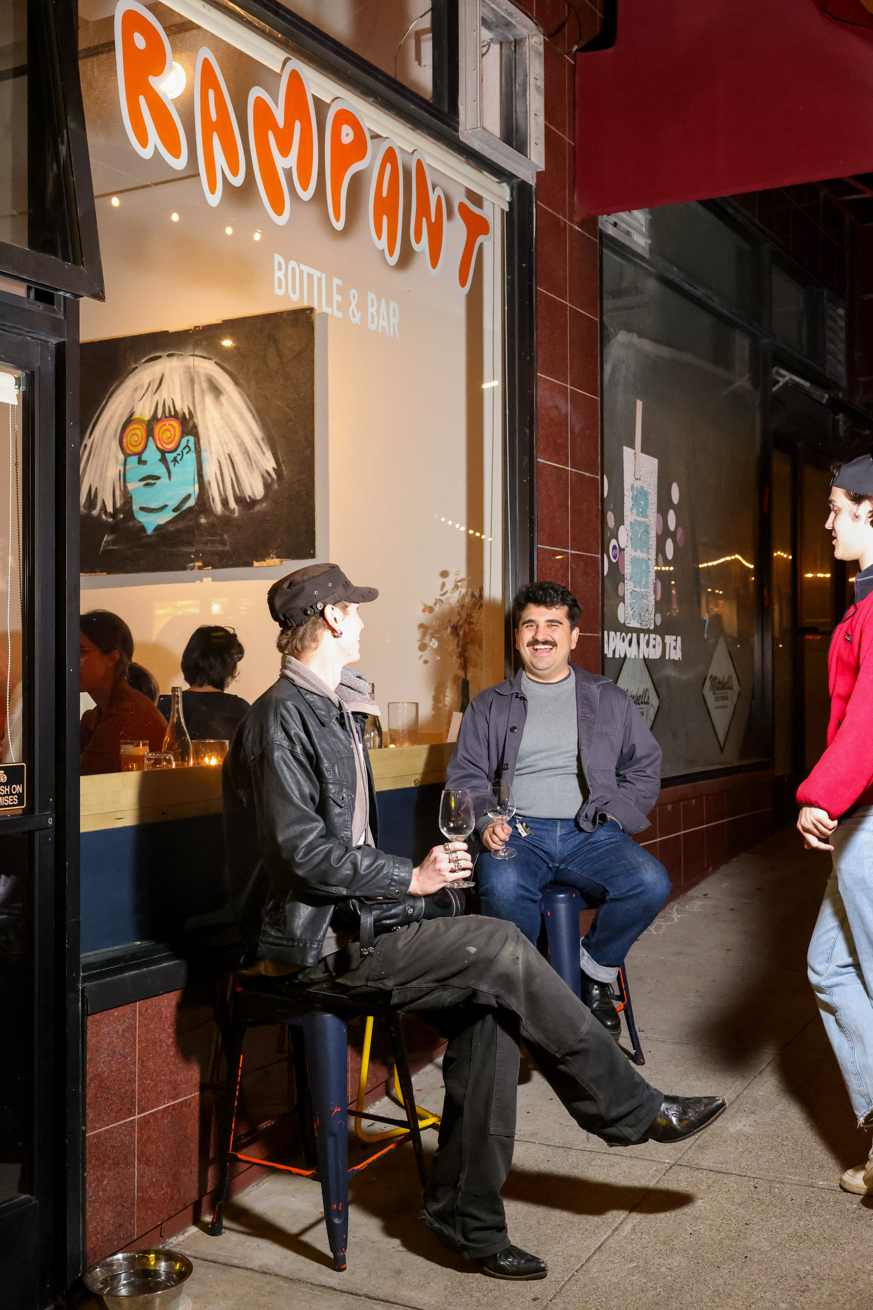 Two people are sitting outside a bar called &quot;Rampant,&quot; holding drinks and smiling, while another person stands nearby. There's modern artwork visible inside.