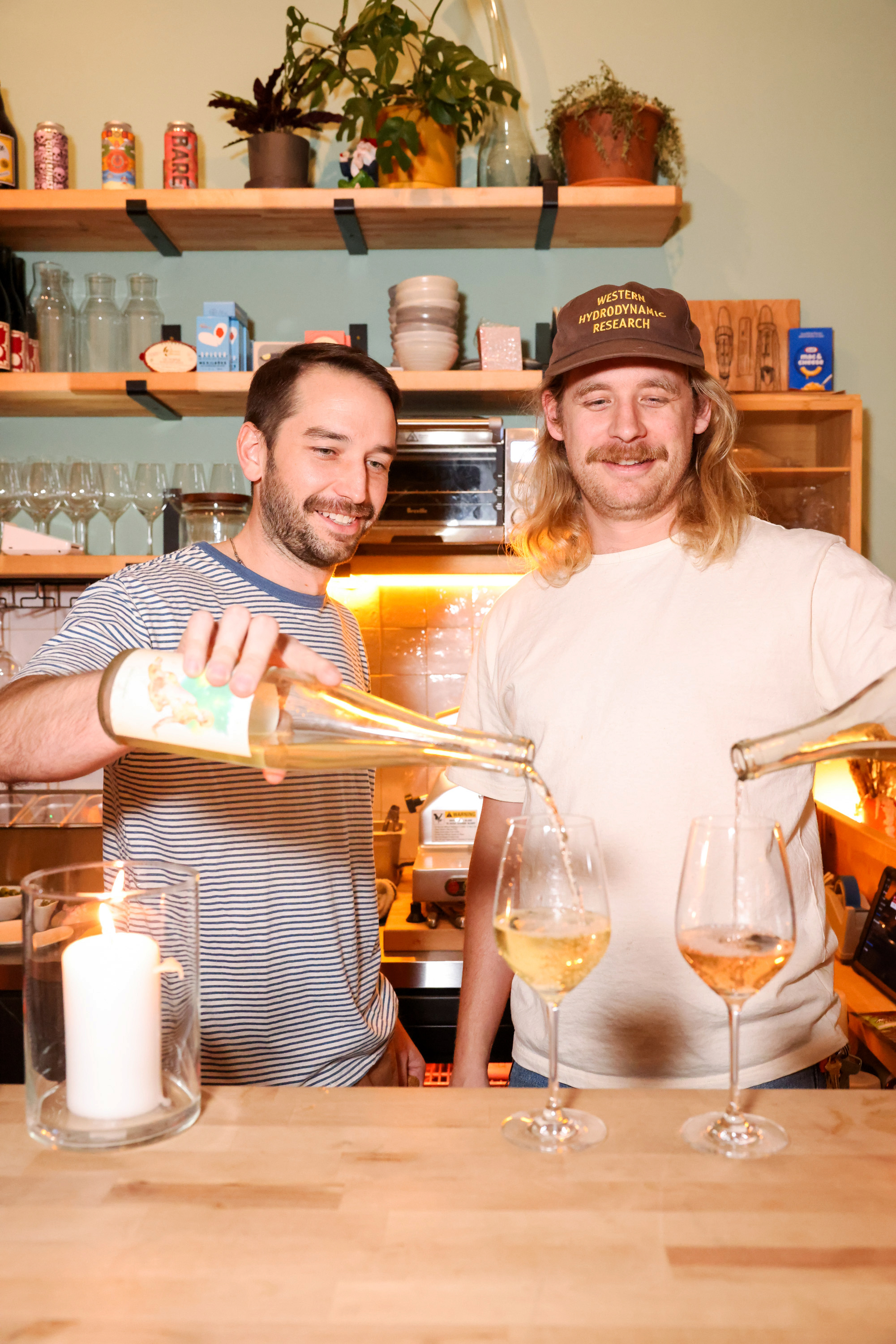 Two people are happily pouring wine into glasses at a wooden counter. A lit candle is nearby, and shelves with various items are in the background.