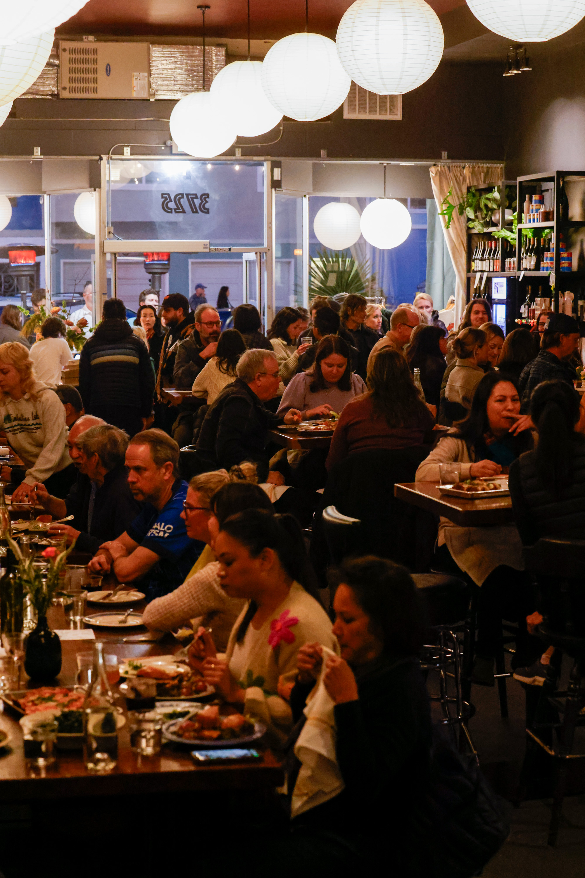 A bustling restaurant, filled with people enjoying their meals. Soft lighting from paper lanterns creates a warm ambiance, and shelves hold various beverages.