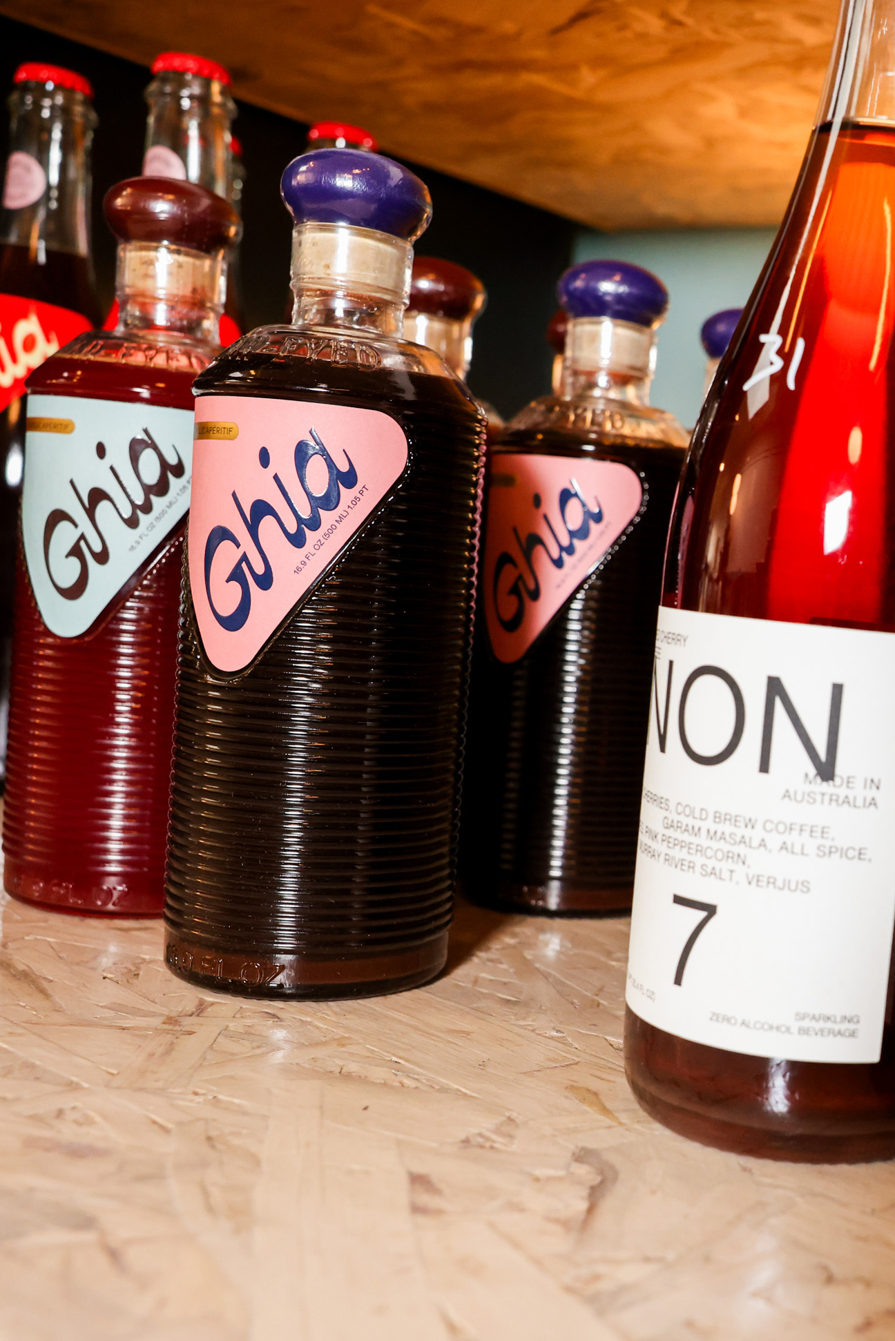 The image shows several bottles of Ghia, a non-alcoholic aperitif with blue caps and pink labels, next to a NON beverage bottle with a white label.