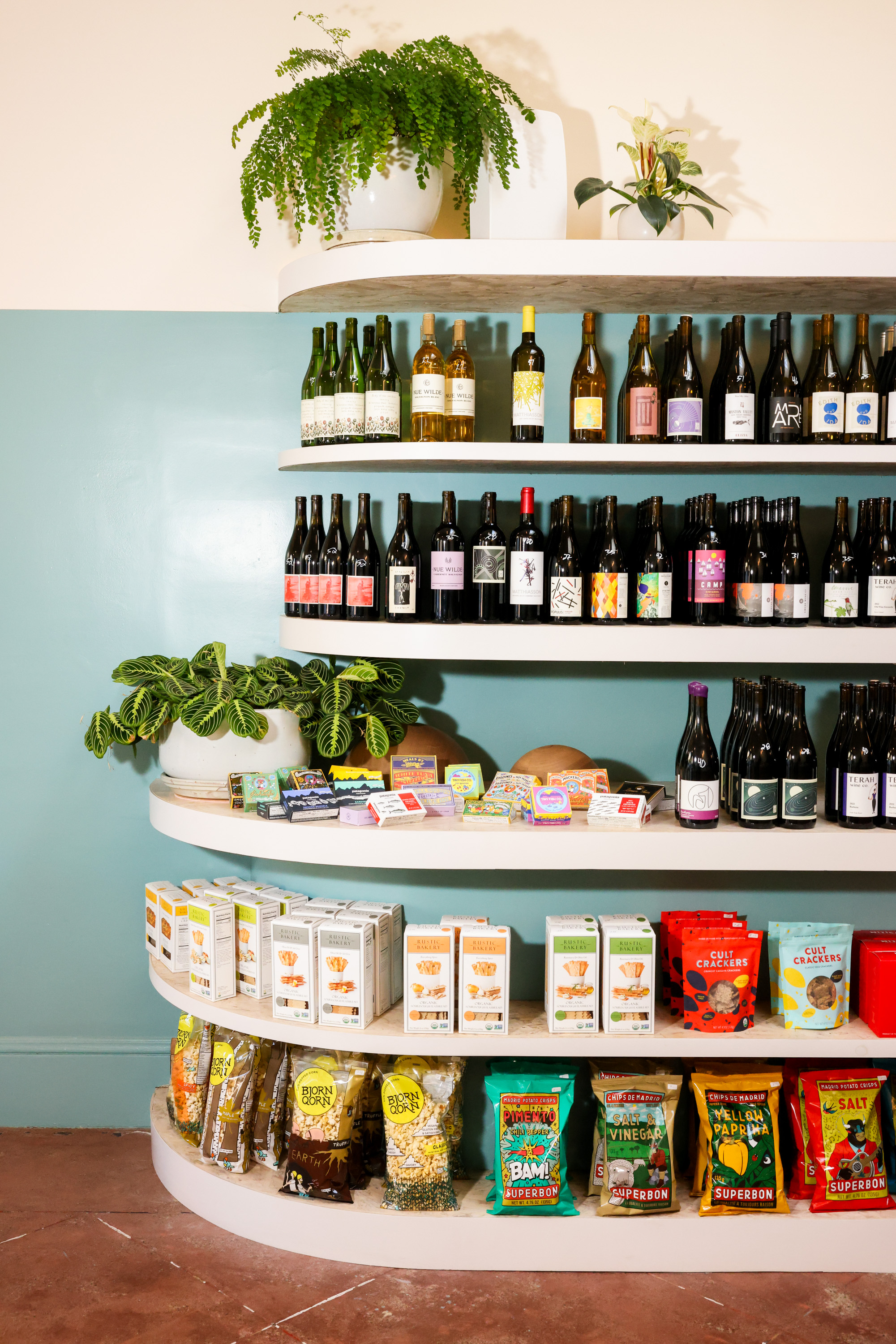 The image shows shelves with potted plants on top, wine bottles in the middle, and snacks and boxed goods on the lower shelves against a light blue wall.